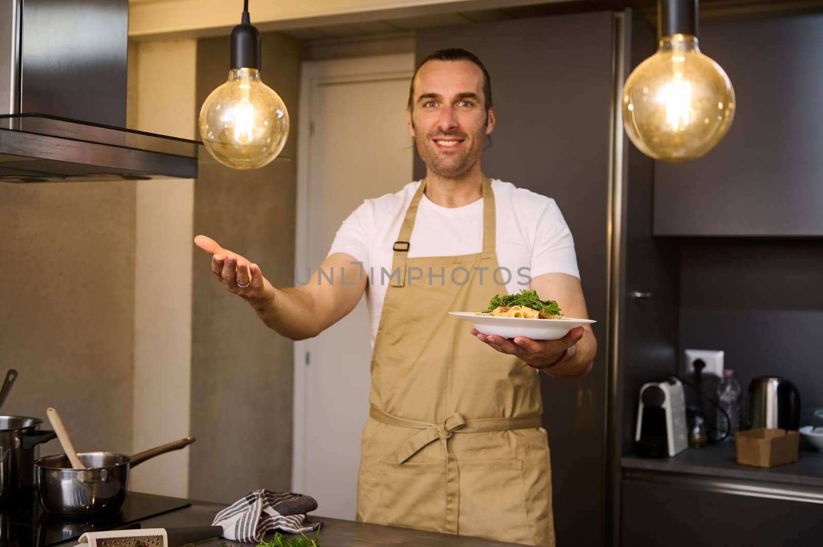 Authentic portrait of a smiling Caucasian man in beige chef's apron, holding a plate with Italian spaghetti pasta and showing it at camera. standing modern home kitchen interior. People and culinary