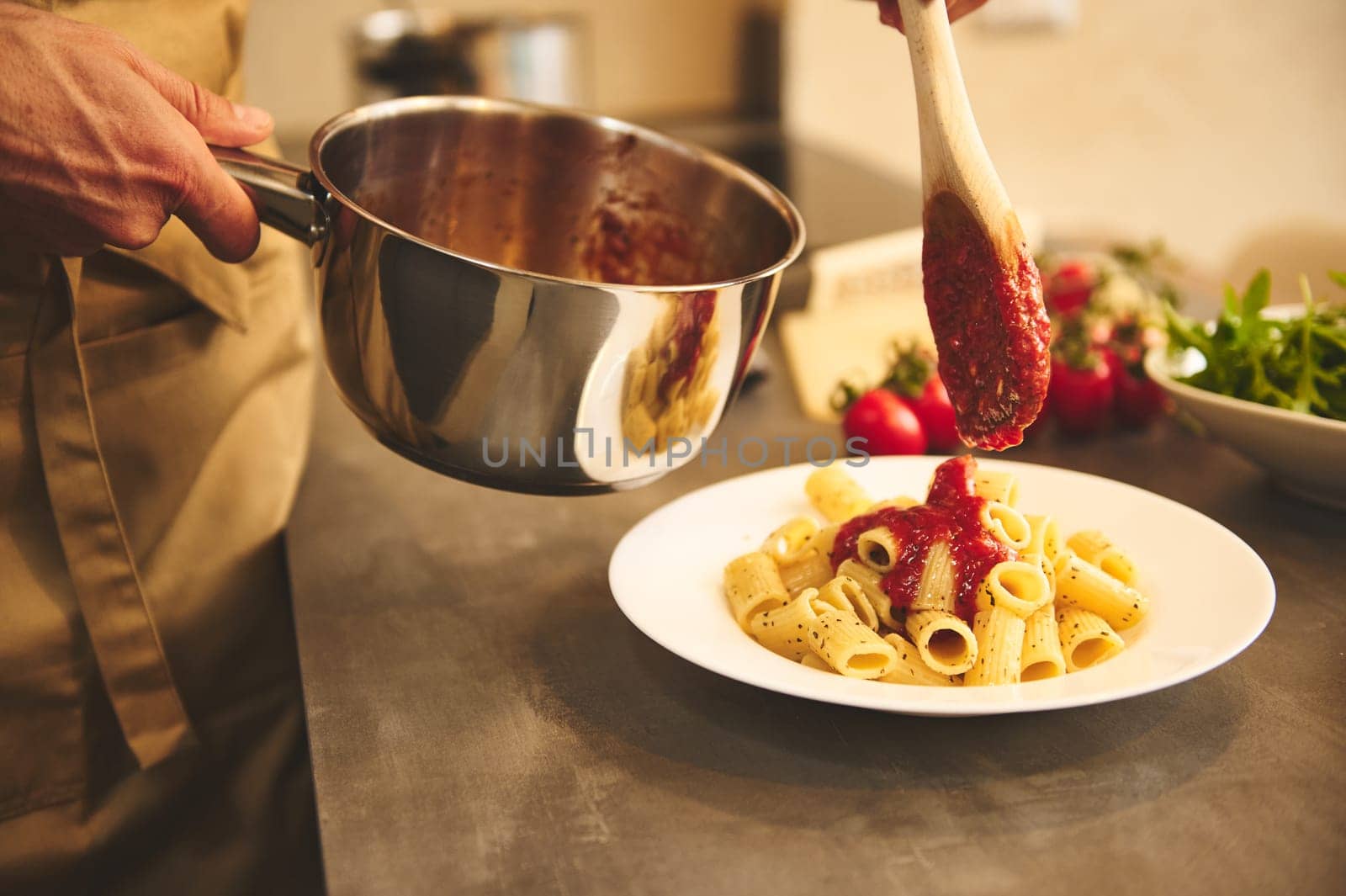 Hands of a man holding a stainless steel pan and pouring tomato sauce on pasta, cooking dinner at modern home kitchen by artgf