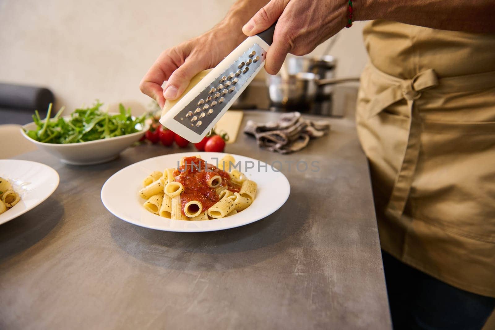 Close-up view of male chef plating up pasta before serving it to the customer. Hands holding grater, grating parmesan cheese, seasoning Italian pasta spaghetti while cooking dinner at home kitchen by artgf
