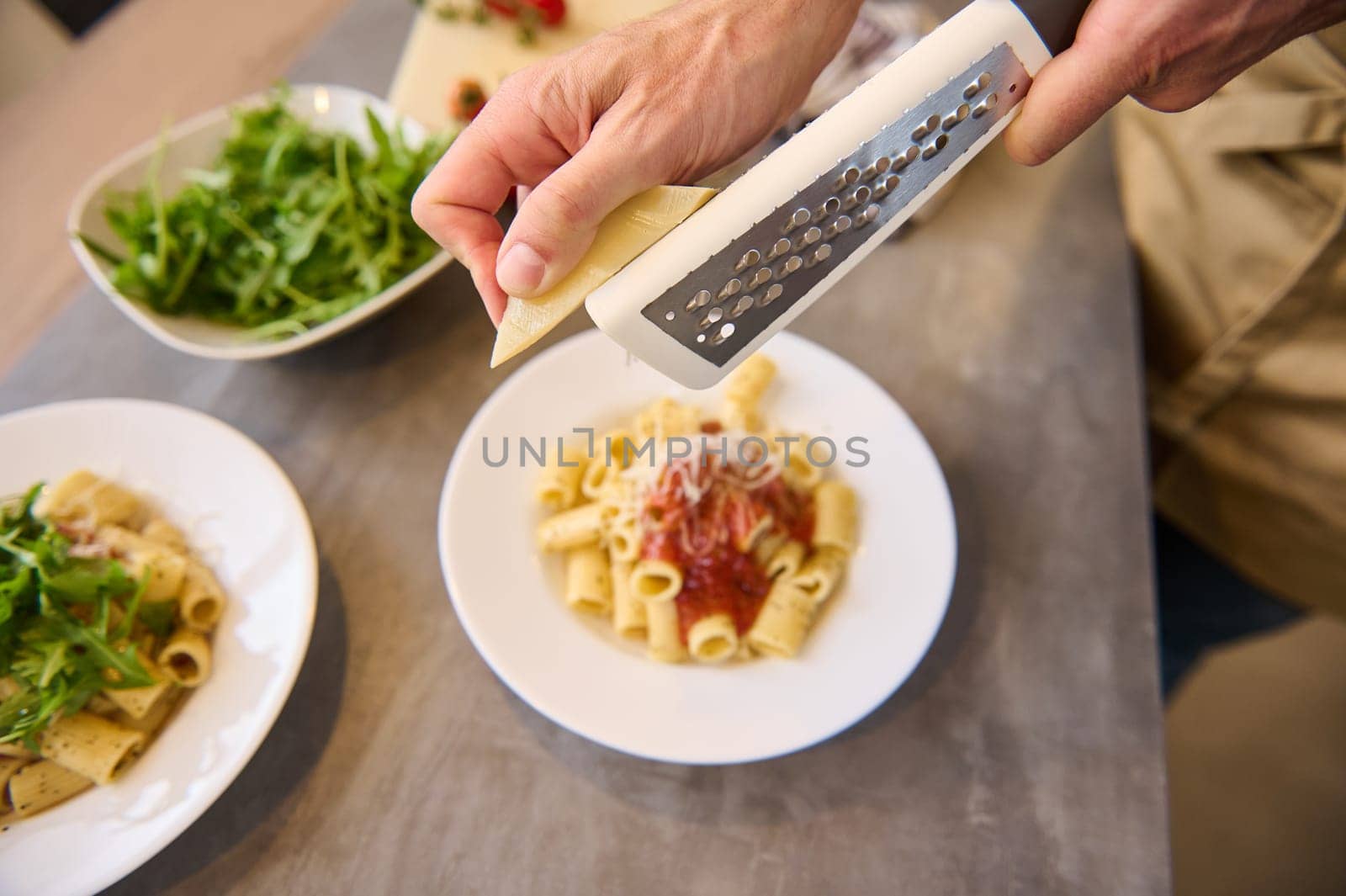 Directly above chef hands using grater, grating cheese on freshly cooked pasta with tomato sauce, seasoning and plating up the meal before serving it by artgf