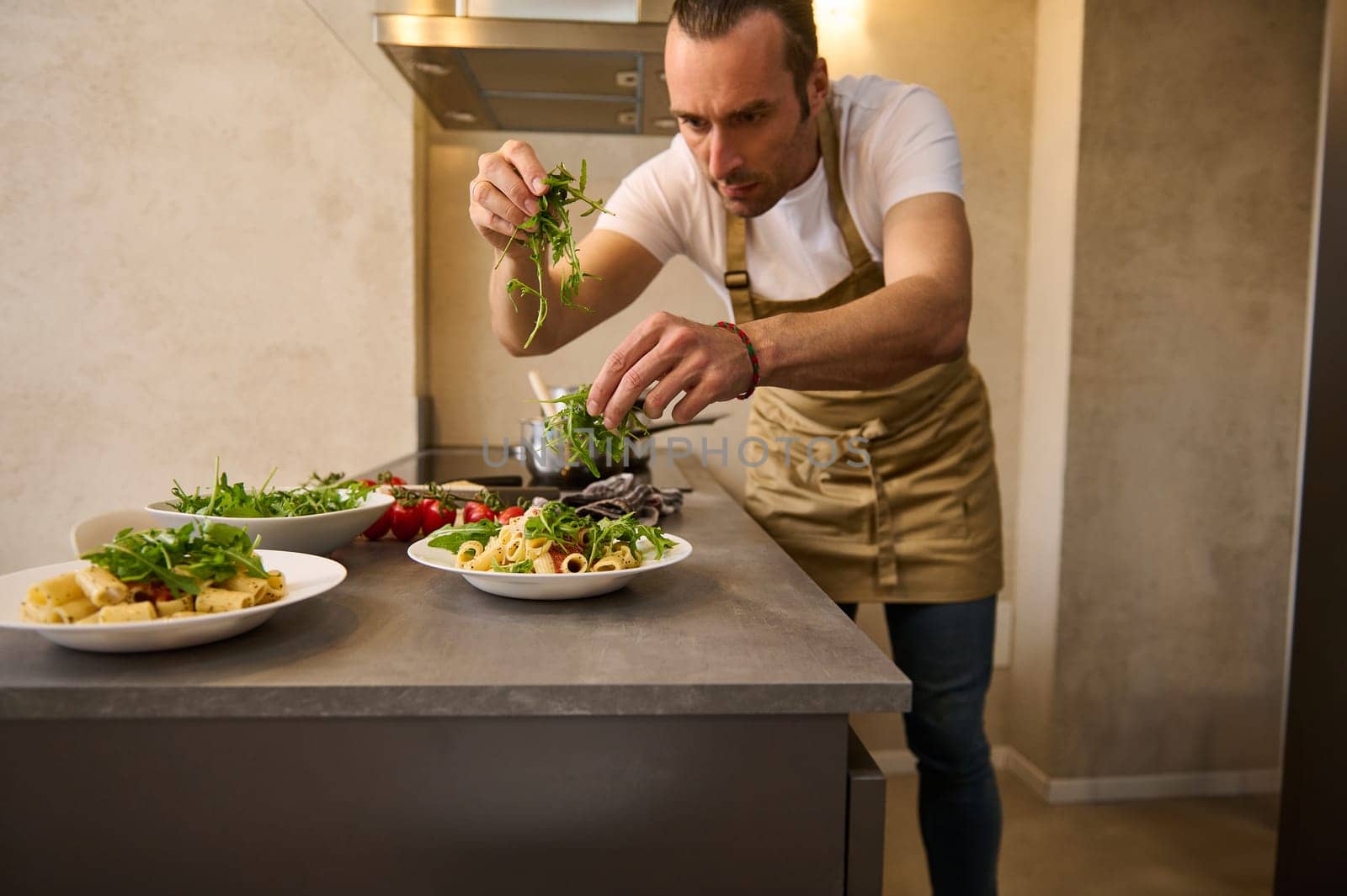 Handsome male chef cooks delicious Italian pasta with the sauce, seasoning with fresh arugula leaves and grated parmesan cheese on the pasta in a plate, standing at kitchen counter. Decoration of dish