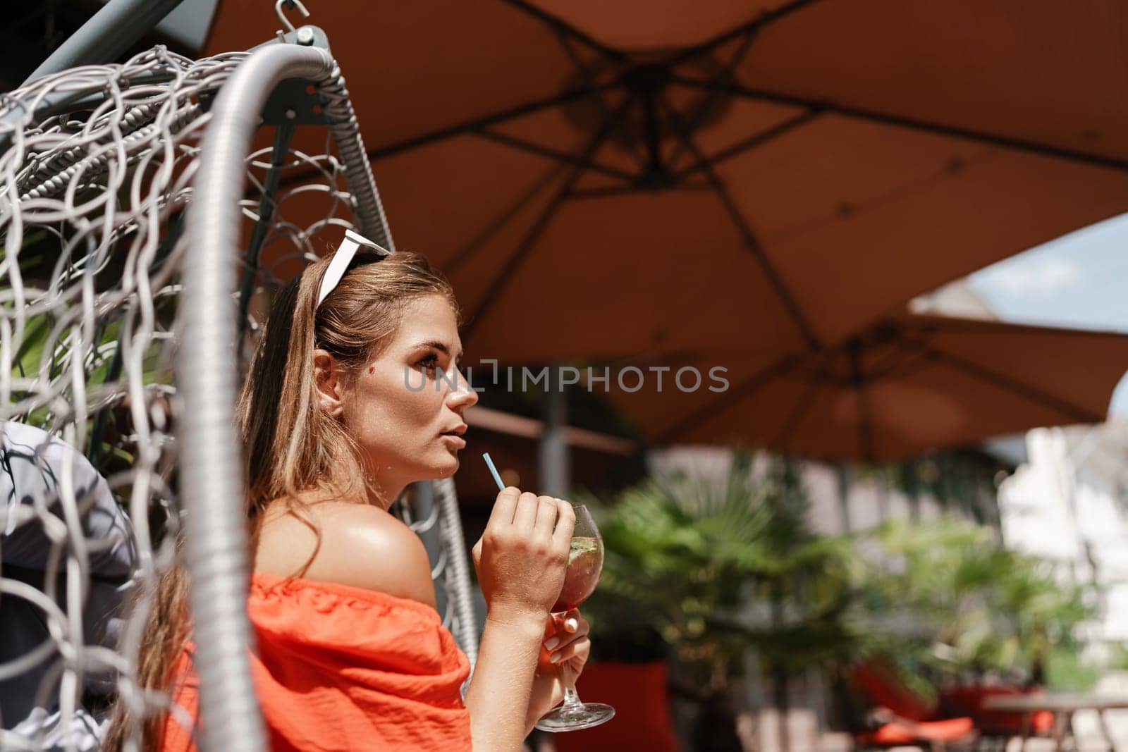 A woman in an orange dress is sitting in a chair with a drink in front of her. She is looking at the camera and she is enjoying her drink. The scene is set in a patio or outdoor area. by Matiunina