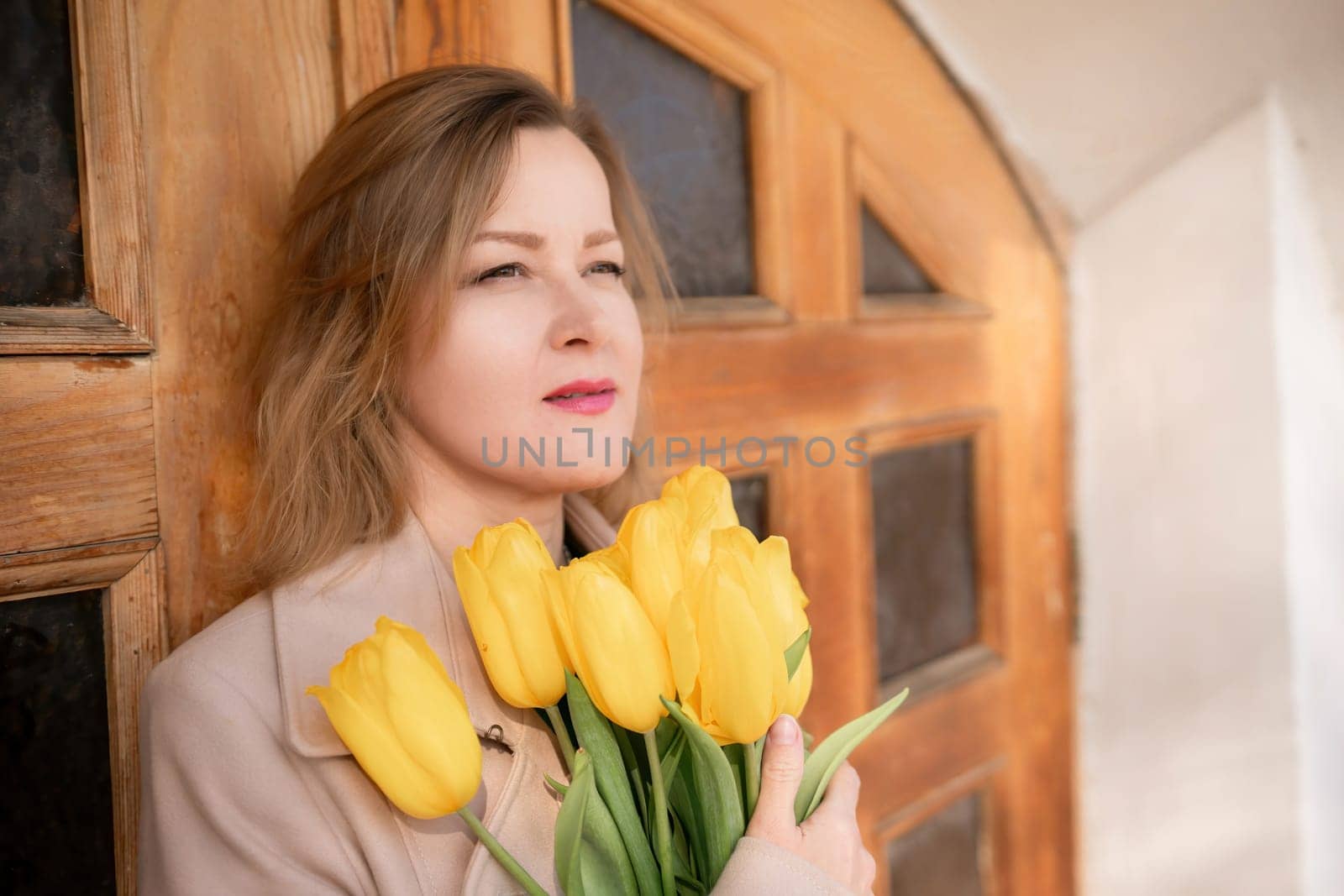 A woman is holding a bouquet of yellow tulips. She is wearing a tan coat and a green shirt