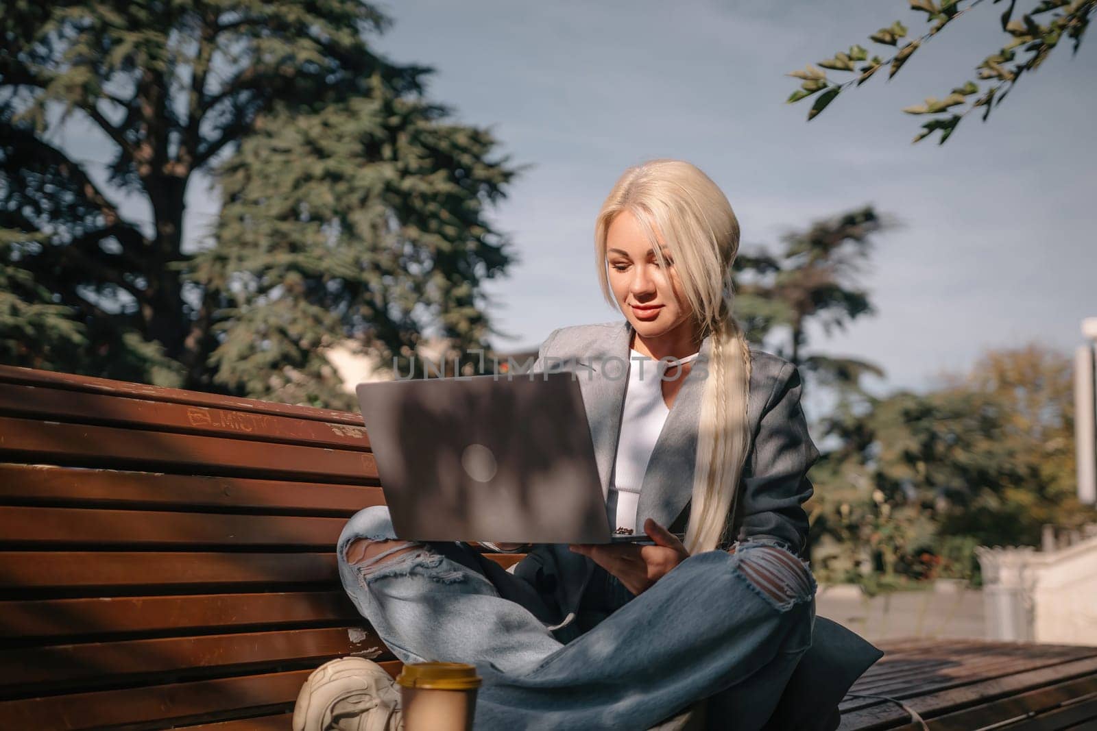 A blonde woman sits on a bench with a laptop in front of her. She is wearing a gray jacket and jeans. The scene suggests a casual and relaxed atmosphere, as the woman is using her laptop outdoors