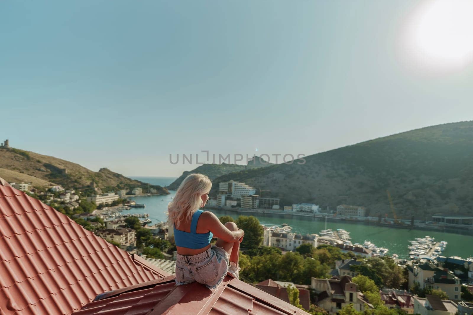 Woman sits on rooftop, enjoys town view and sea mountains. Peaceful rooftop relaxation. Below her, there is a town with several boats visible in the water. Rooftop vantage point. by Matiunina