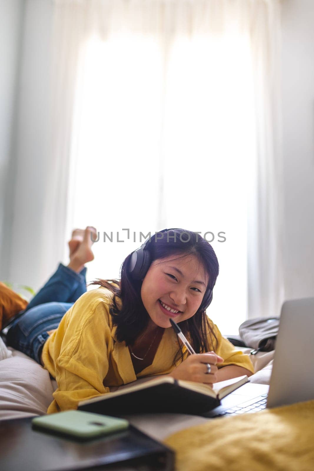 Happy Chinese teen girl student doing homework at home bedroom lying on bed. Chinese teenager studying at home with laptop looking at camera. Vertical. Education concept.