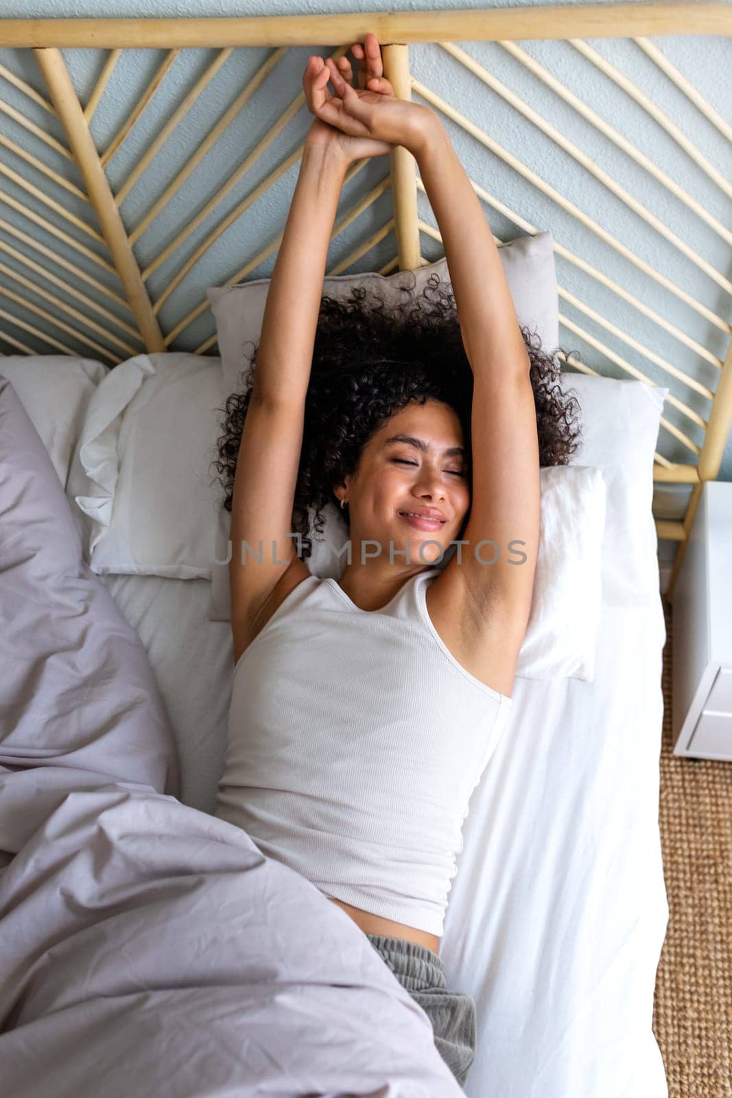 Young African American woman waking up in the morning, stretching arms in bed. Good morning. Vertical image. Lifestyle concept.