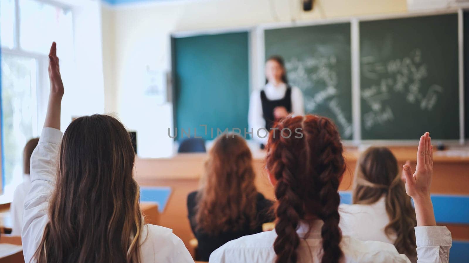 Intelligent group of young school children all raising their hands in the air to answer a question posed by the female teacher, view from behind