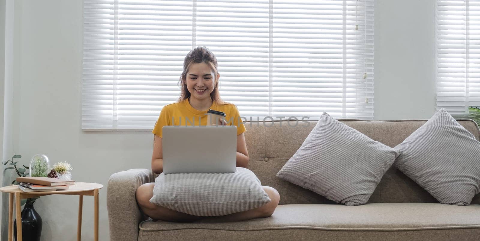 A woman is sitting on a couch with a laptop in front of her. She is smiling and holding a cup. Concept of relaxation and leisure