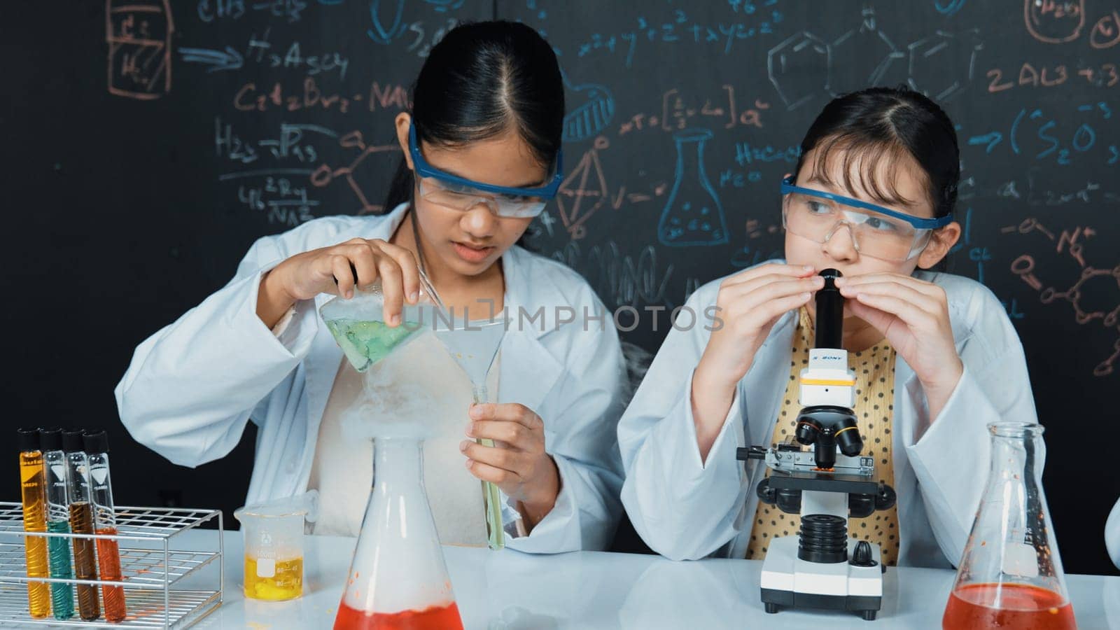 Young highschool student doing experiment by pouring sample in test tube. Girl wearing glasses and looking under microscope to inspect sample in STEM science class. Creative education. Edification.
