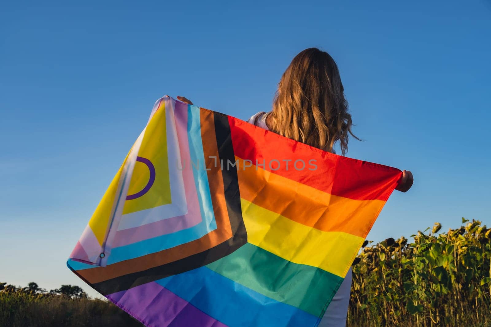 Symbol of LGBTQ pride month. Young woman showing Rainbow LGBTQIA flag waving in wind made from silk material on field background. Equal rights by anna_stasiia