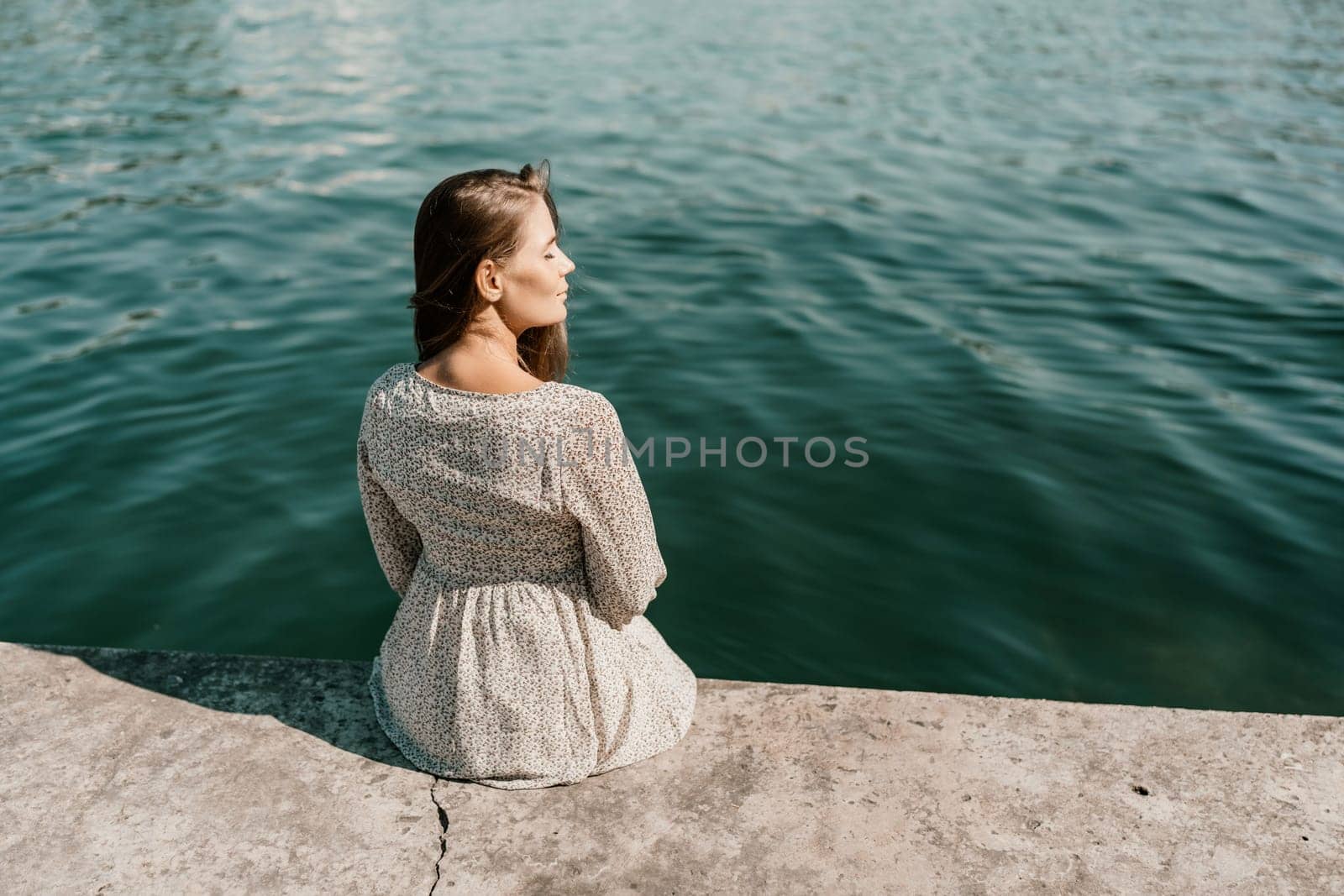 A woman is sitting on a ledge by a body of water, wearing a dress with a leopard print pattern. The scene is peaceful and serene, with the woman enjoying the view of the water and the surrounding area