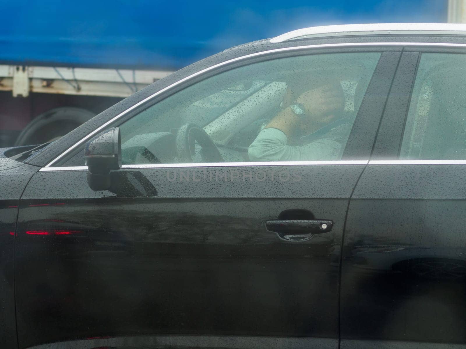 A person is sleeping in the driver's seat of a black car. The car is parked in a parking lot