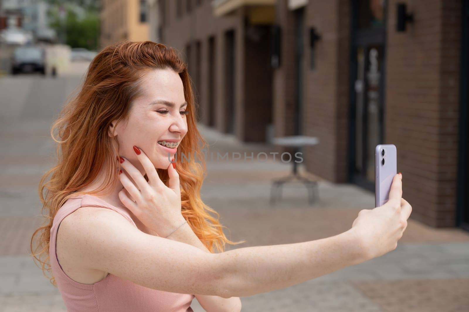 Young woman with braces on her teeth smiles and takes a selfie on a smartphone outdoors