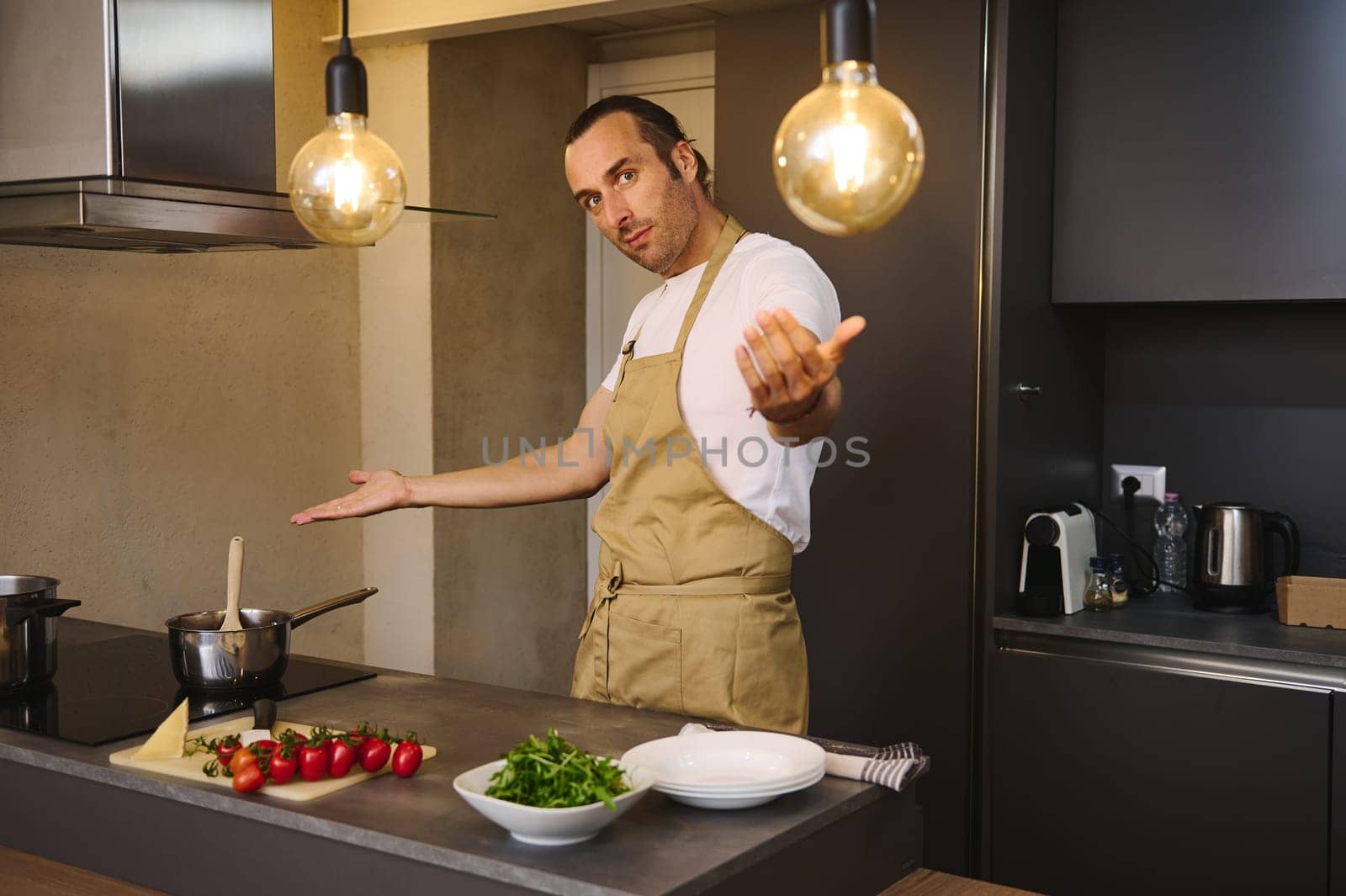 Handsome Caucasian man in chef's apron cooking family dinner, smiling and showing the kitchen counter with fresh ingredients looking at camera by artgf