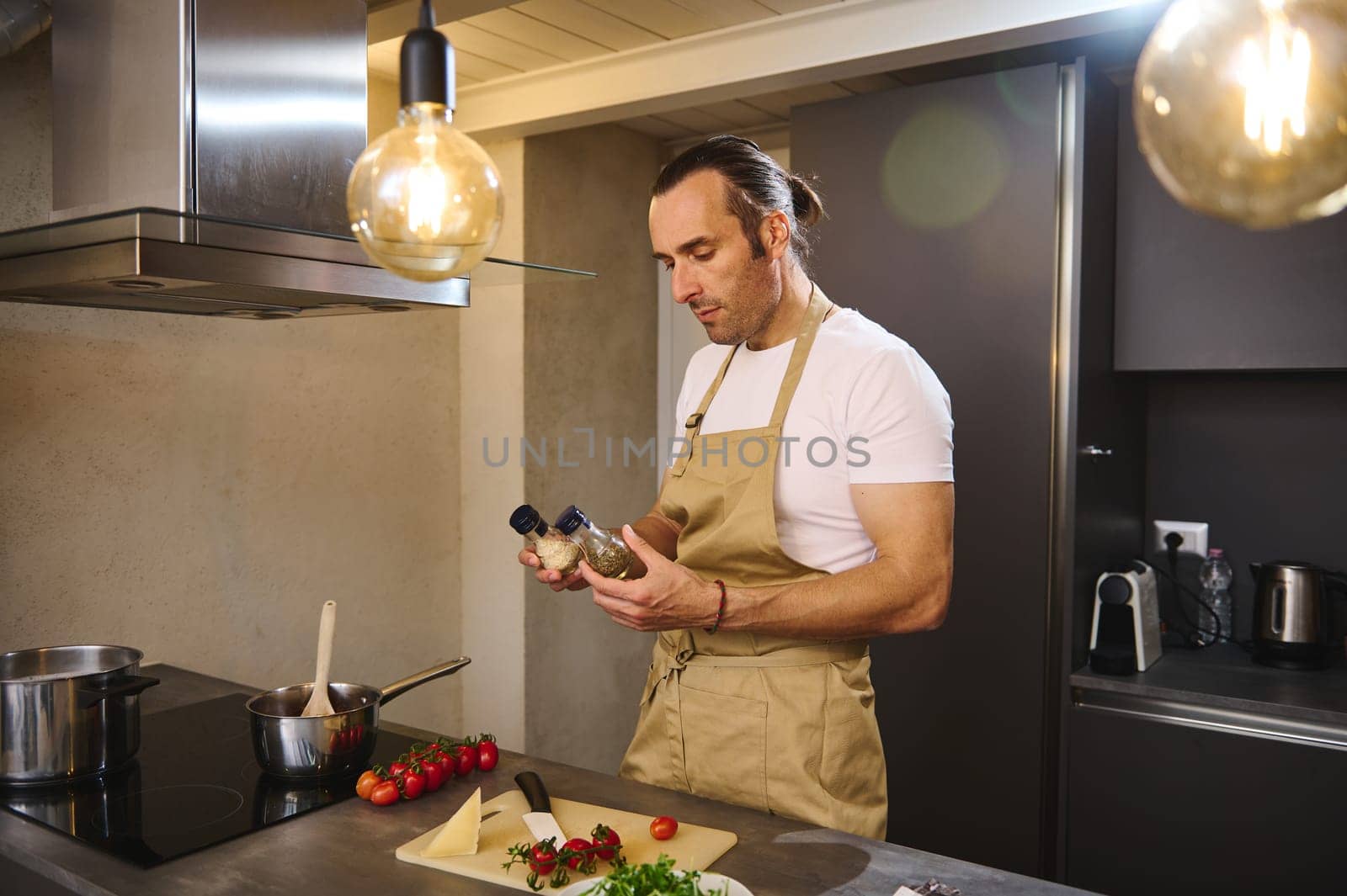 Handsome European male chef in beige kitchen apron, holding bottles of condiments for seasoning dish, standing at kitchen counter with fresh ingredients, cooking dinner at home kitchen