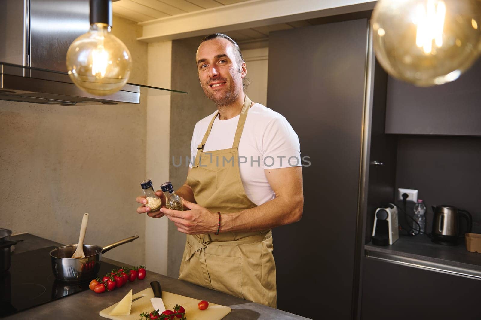 Portrait of male chef in beige kitchen apron, holding bottles of condiments for seasoning dish, standing at kitchen counter with fresh ingredients, looking at camera, preparing dinner at home kitchen