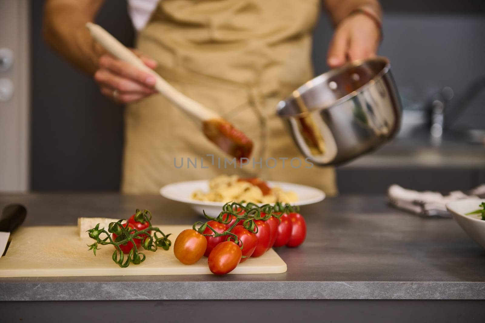 A branch of fresh ripe organic tomato cherry on the cutting board against the backdrop of a blurred male chef cooking Italian pasta at home kitchen. Traditional Italian cuisine. Food background by artgf