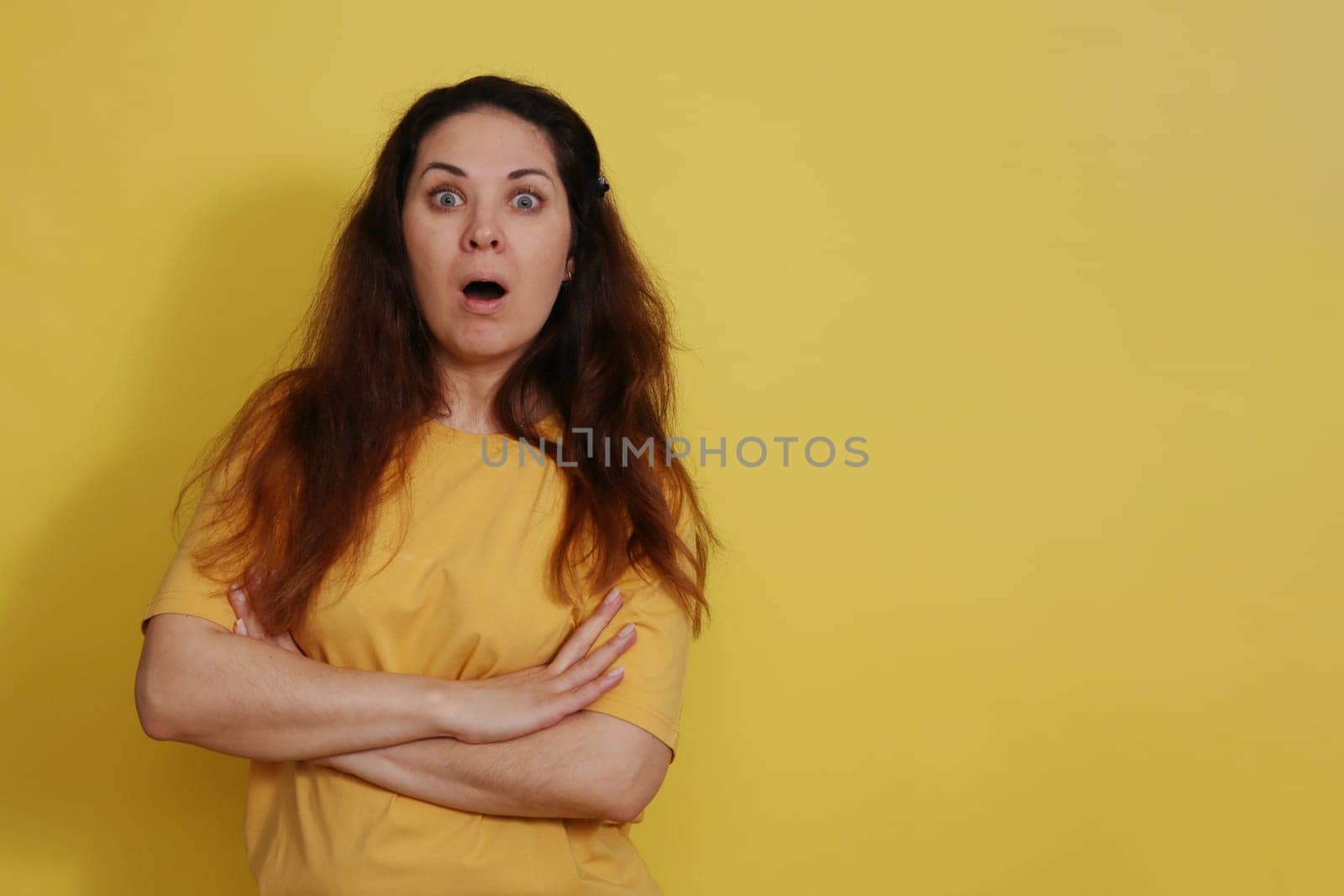 Beautiful woman in yellow t-shirt on yellow studio background with long dark hair opened her mouth in surprise. Portrait of a surprised beautiful woman with the expectation of darkness in a yellow tank top on a yellow background.