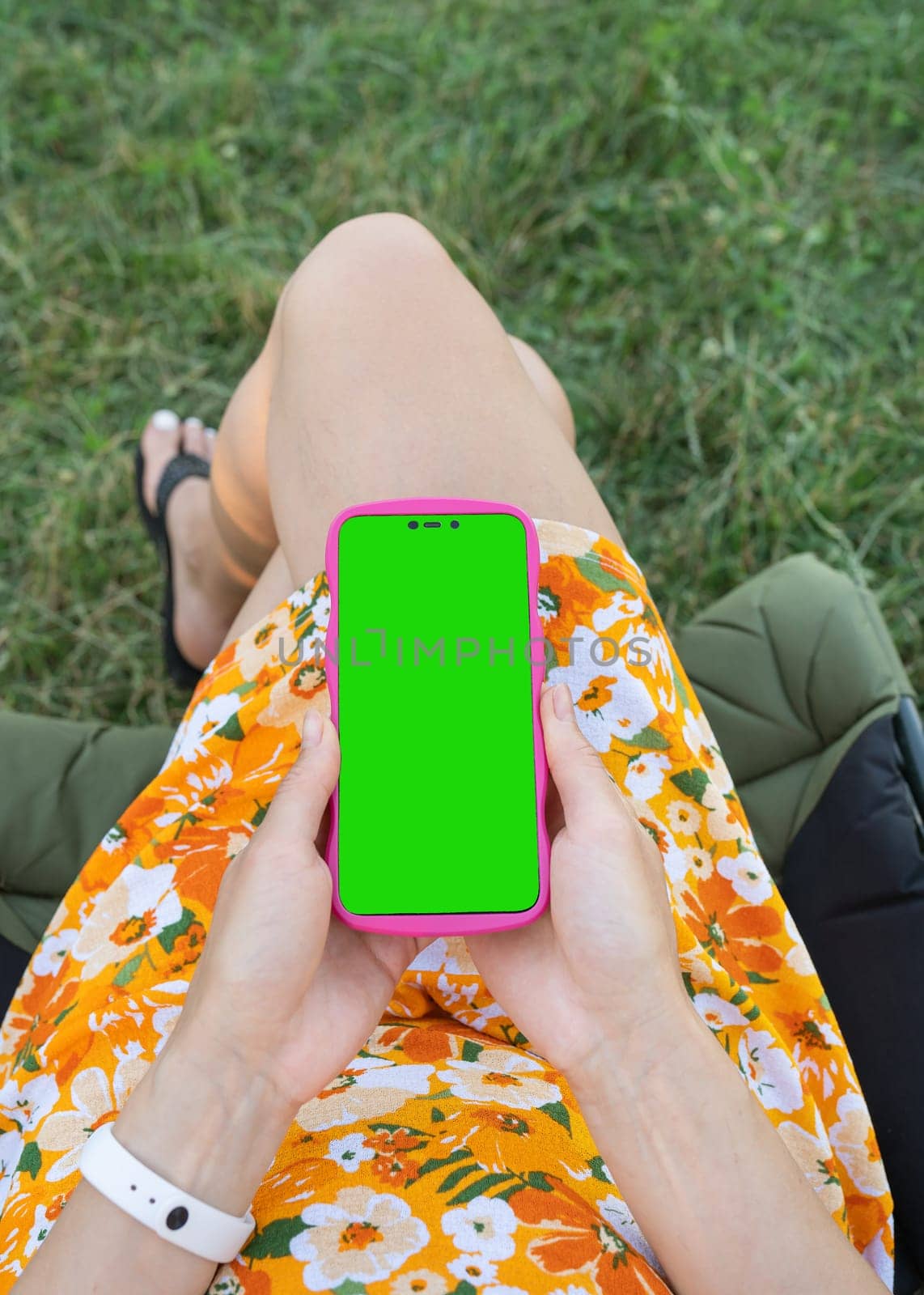 A girl in a floral dress holds a smartphone with a green screen and sits on the grass. Template, place for inscription