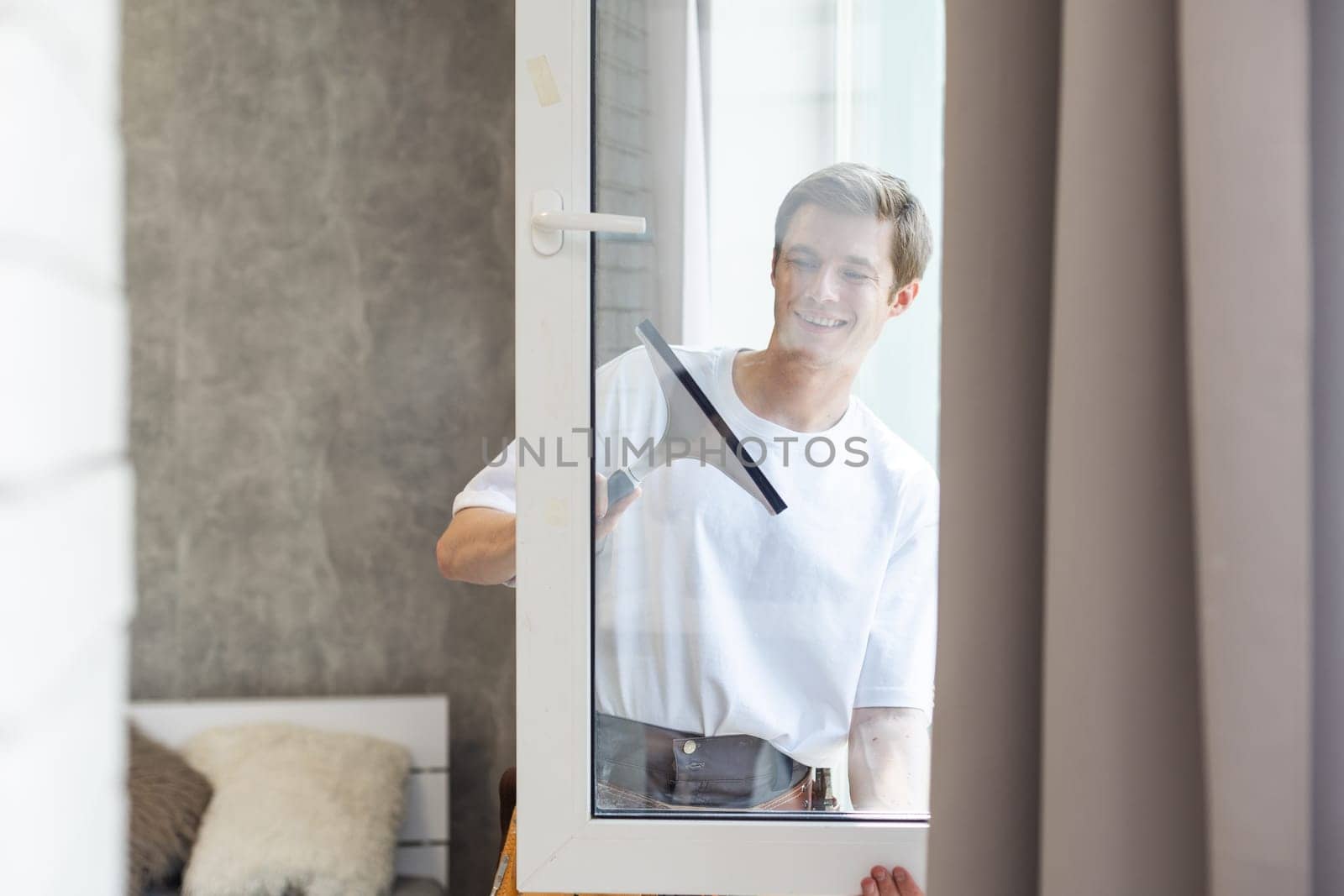 Side view of smiling man with bottle of detergent and rag cleaning window at home.