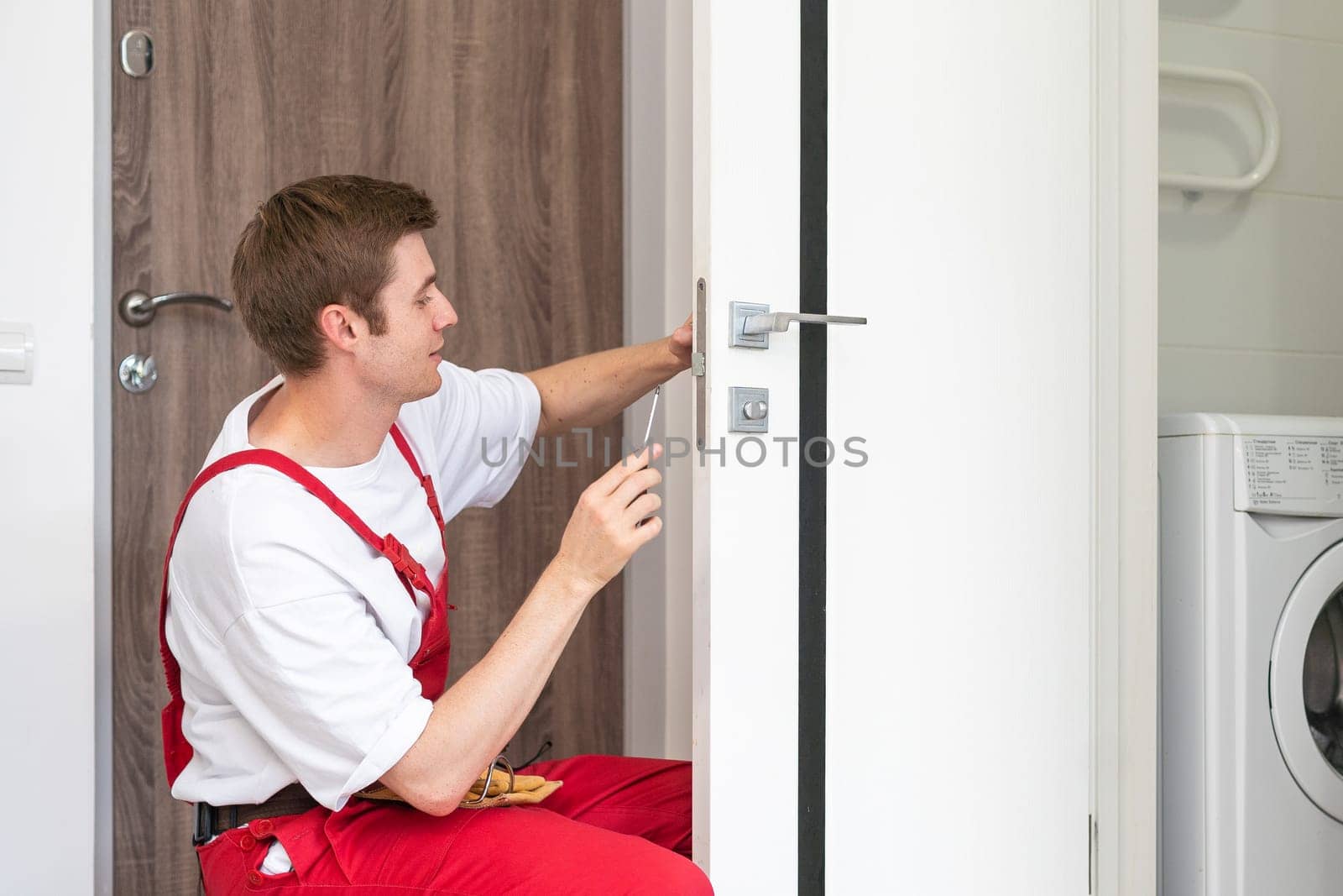 Young handyman installing a white door with an electric hand drill in a room.