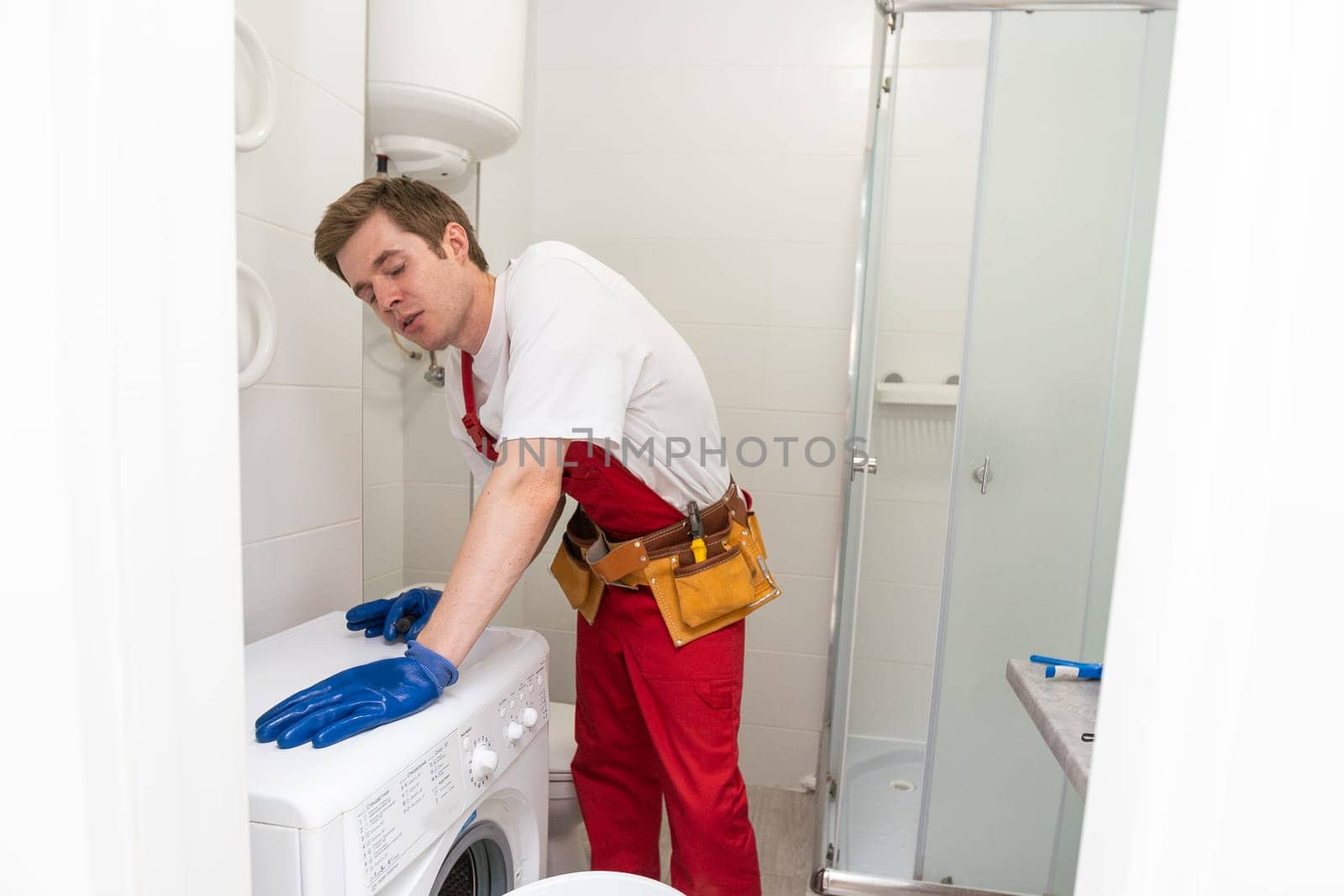The young handsome repairman in worker suit with the professional tools box is fixing the washing machine in the bathroom