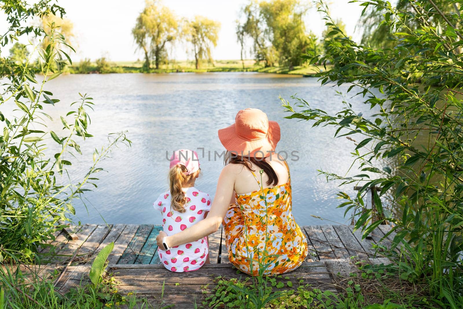 Mother and daughter sit by a serene lake surrounded by lush greenery, evoking a sense of peace and connection with nature. by sfinks