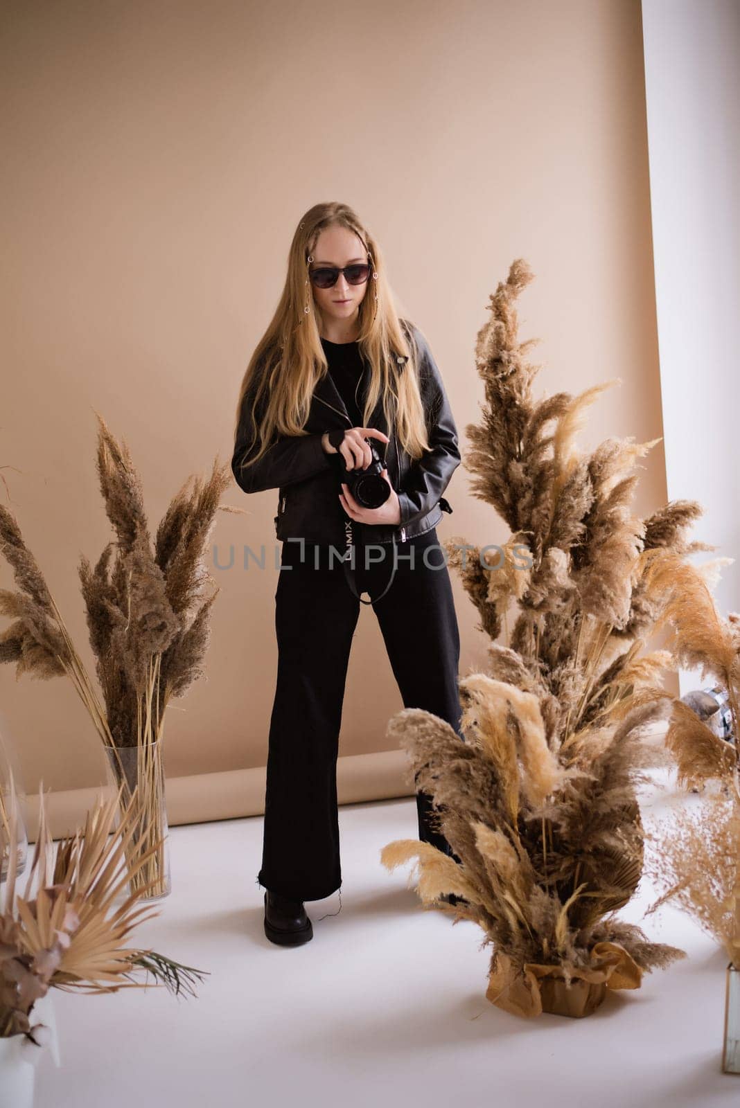 A woman director photographer, a blonde in sunglasses with a camera, seriously in the production studio. Wearing a black clothes on a brown background