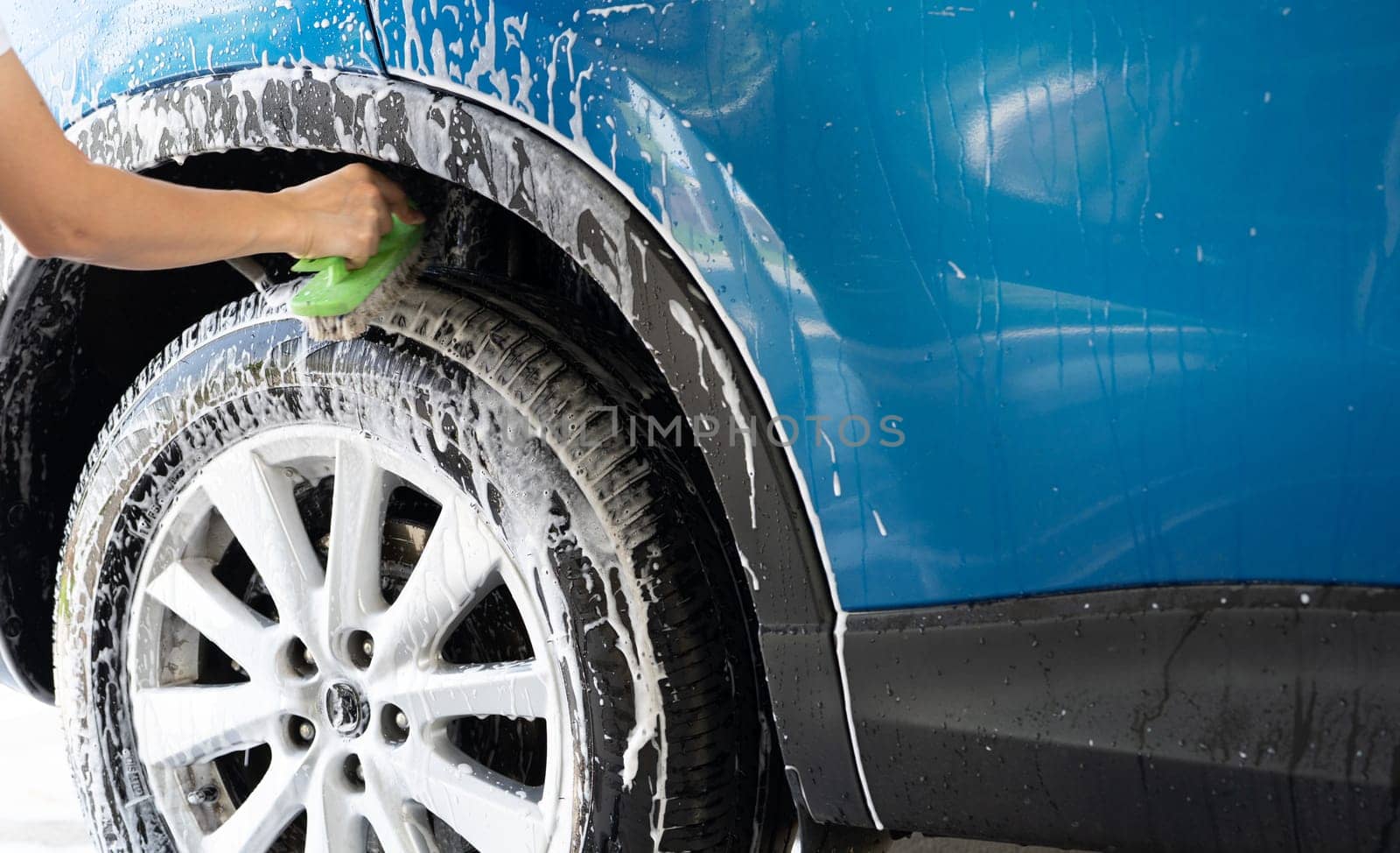 A person is washing a car tire with a green brush. Tire is covered in water and soap. Blue car wash with white soap foam. Auto care service. Car cleaning service concept. Vehicle cleaning service.
