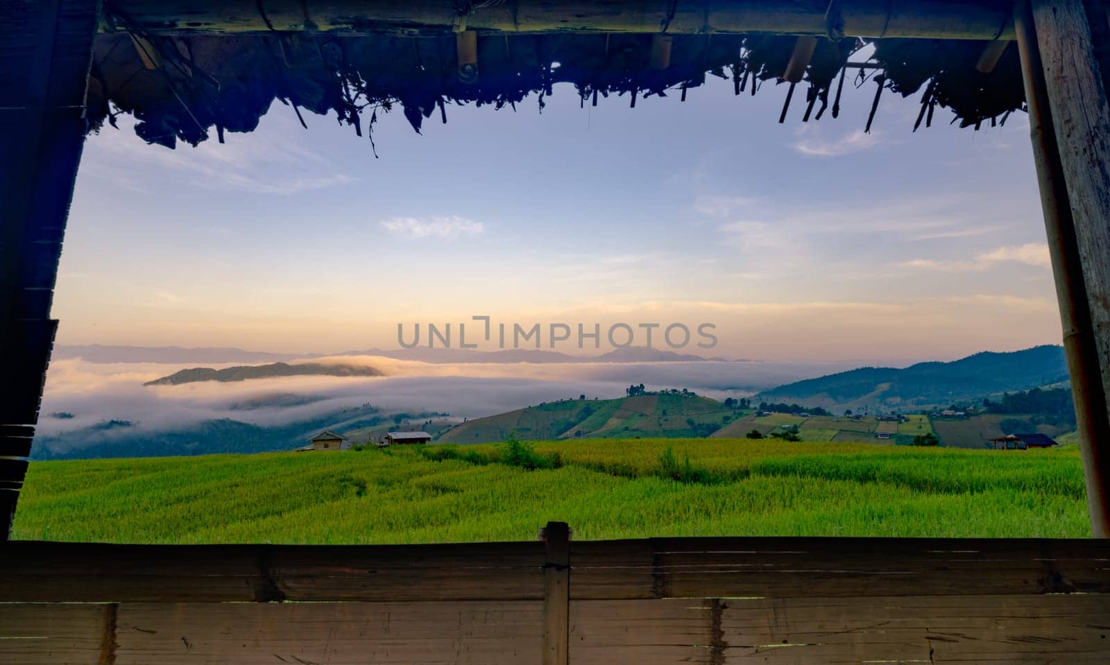 A view of a field with a foggy sky and mountains in the background. The sky is a mix of blue and orange hues, creating a serene and peaceful atmosphere. Rice terrace farm field and mountains. by Fahroni