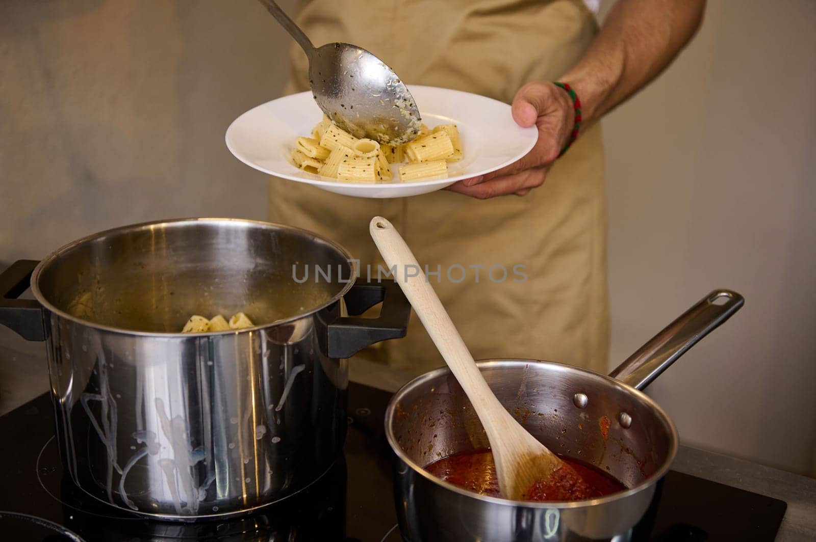 Close-up chef's hands plating up freshly cooked pasta, putting boiled penne on a white plate, standing at induction electric stove with saucepan full of tomato pasta. Italian culinary. Food background