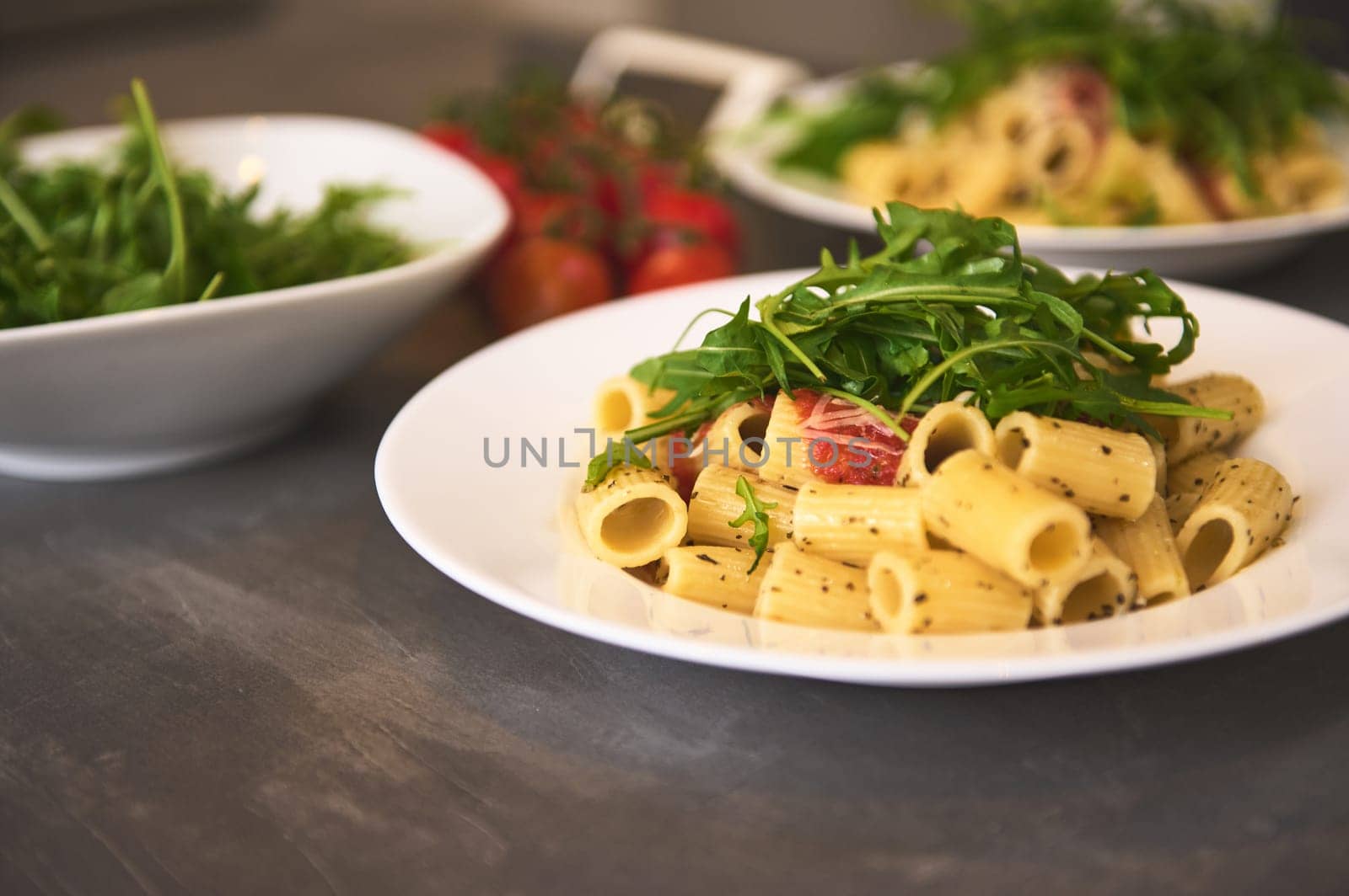 Homemade Italian penne pasta with sauce, tomatoes, arugula, basil and parmesan. Traditional Italian cuisine. Served on a dark table with a branch of organic cherry tomatoes on blurred background. by artgf