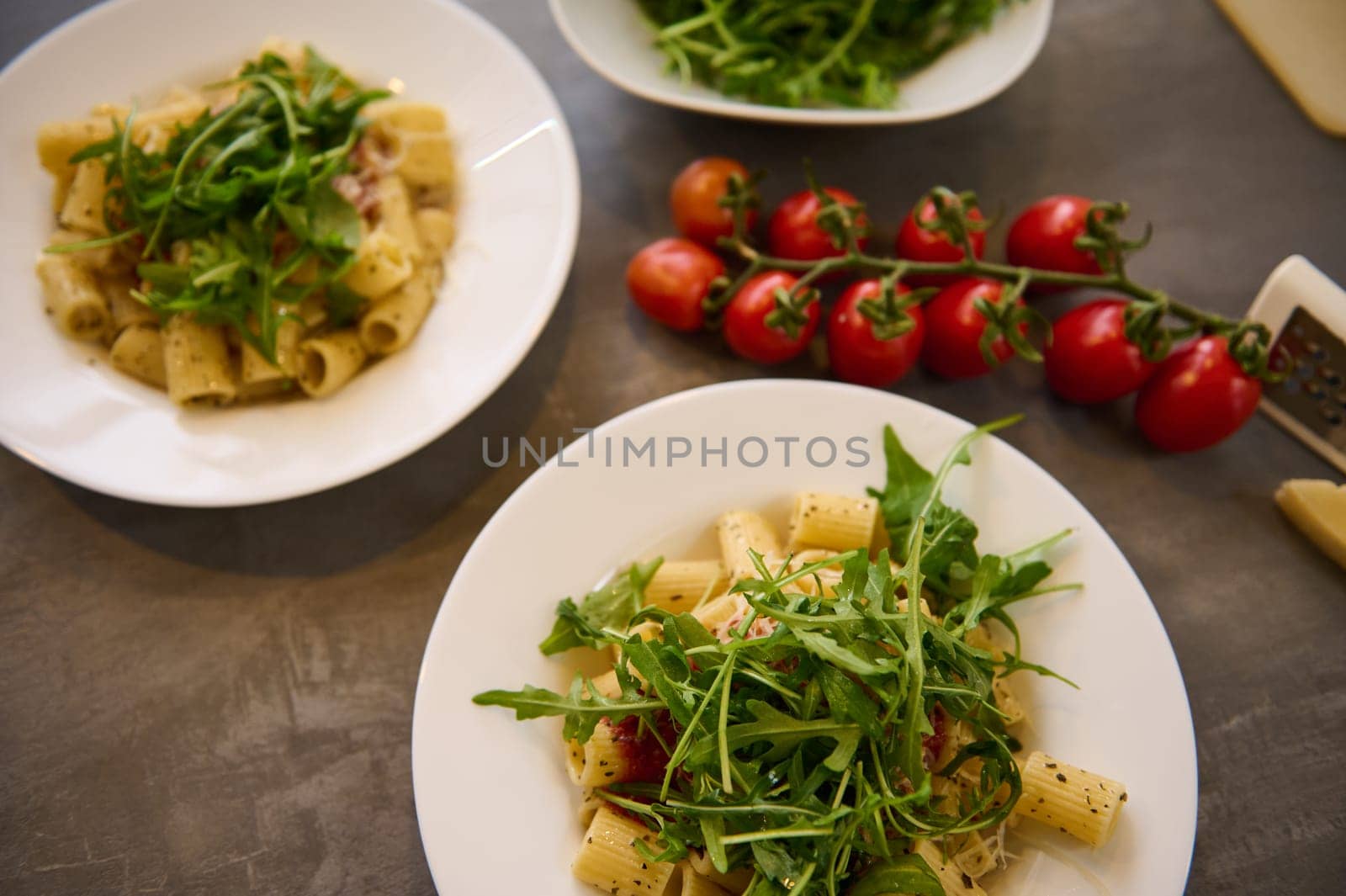 Flat lay white plates of pasta penne with tomato sauce and parmesan cheese, garnished with fresh green arugula leaves on dark background with a branch of cherry tomatoes. Classic Italian cuisine dish.