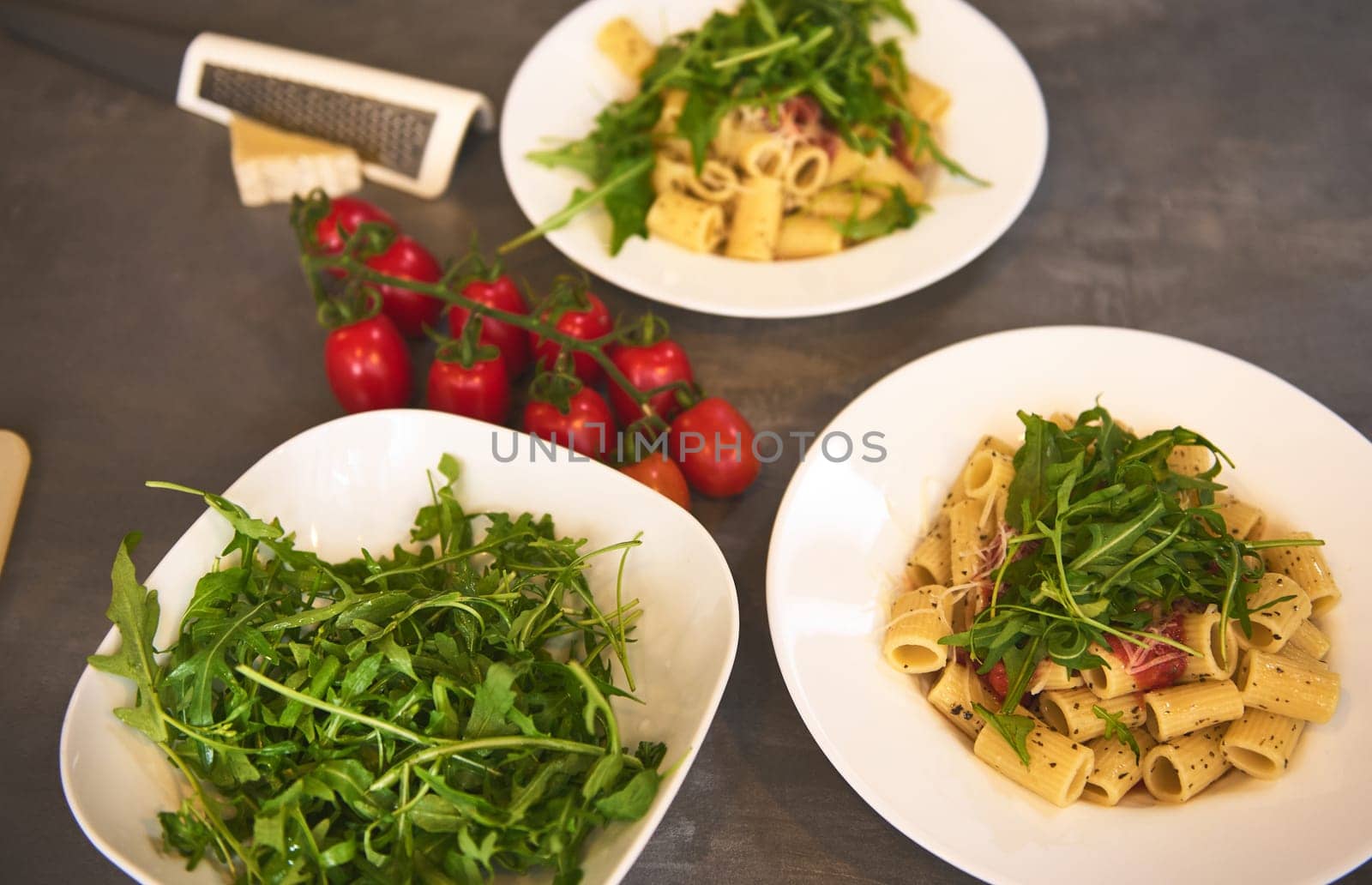 Italian pasta penne with tomato sauce and basil on white plates and bowl of fresh green arugula leaves on a marble background. Food concept by artgf