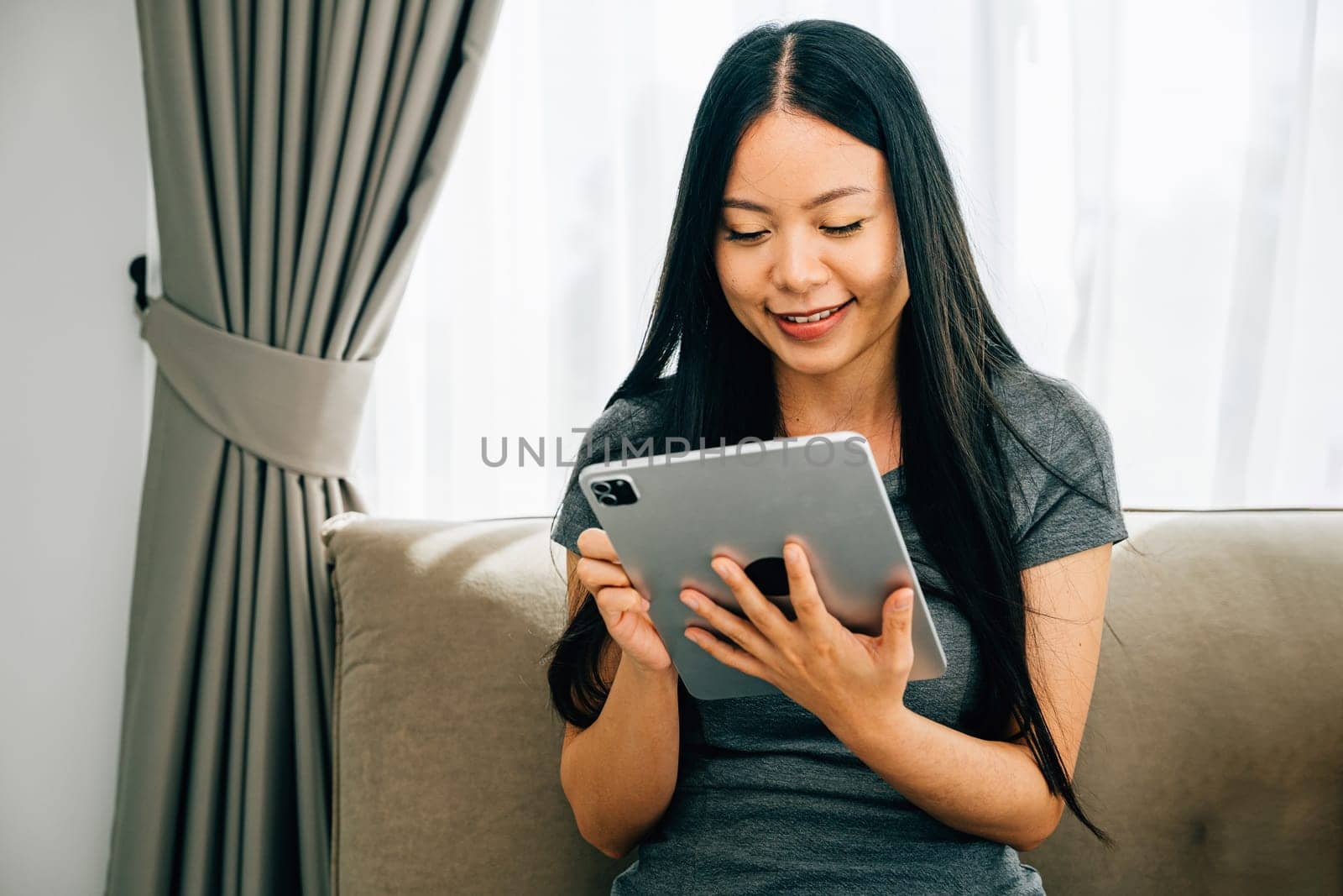 Amidst a comfortable home atmosphere a blissful young Asian woman enjoys leisure time engaged in online shopping through a digital tablet on the sofa exuding relaxation and happiness.