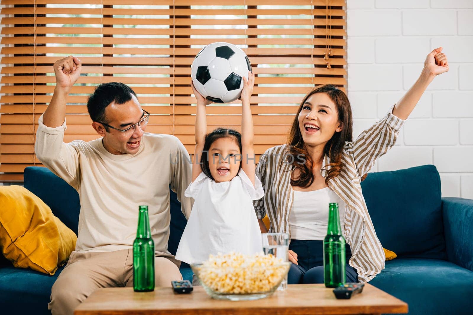 Delighted parents and children, armed with popcorn and a ball, raise their arms and shout in excitement while watching a football match. Their togetherness and bonding celebrate a moment of triumph.