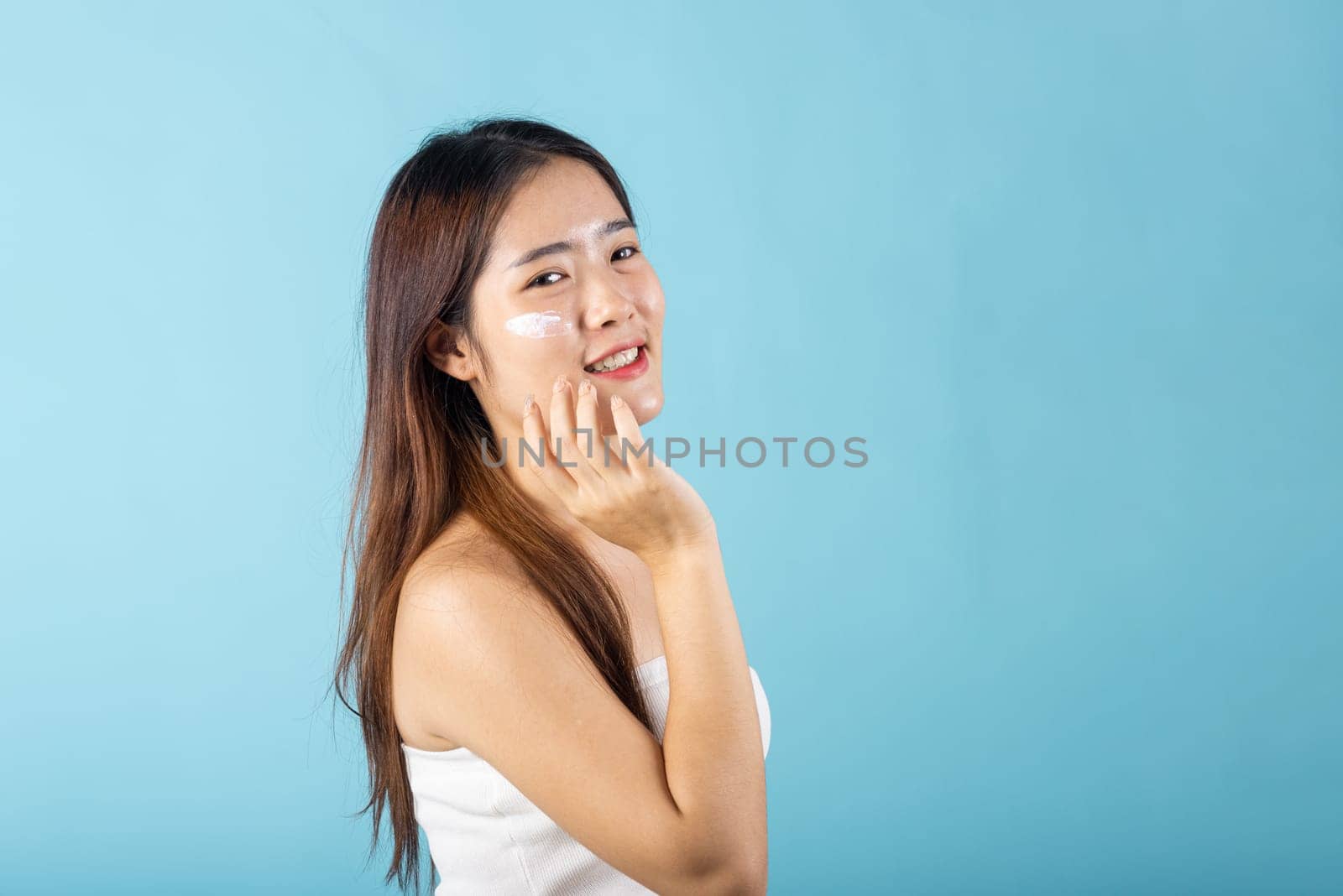 Happy beauty portrait woman gets cream on her cheek, Beautiful young female smiling applying cosmetic cream on her face isolated on blue background, health clean skin, Skin care and beauty concept