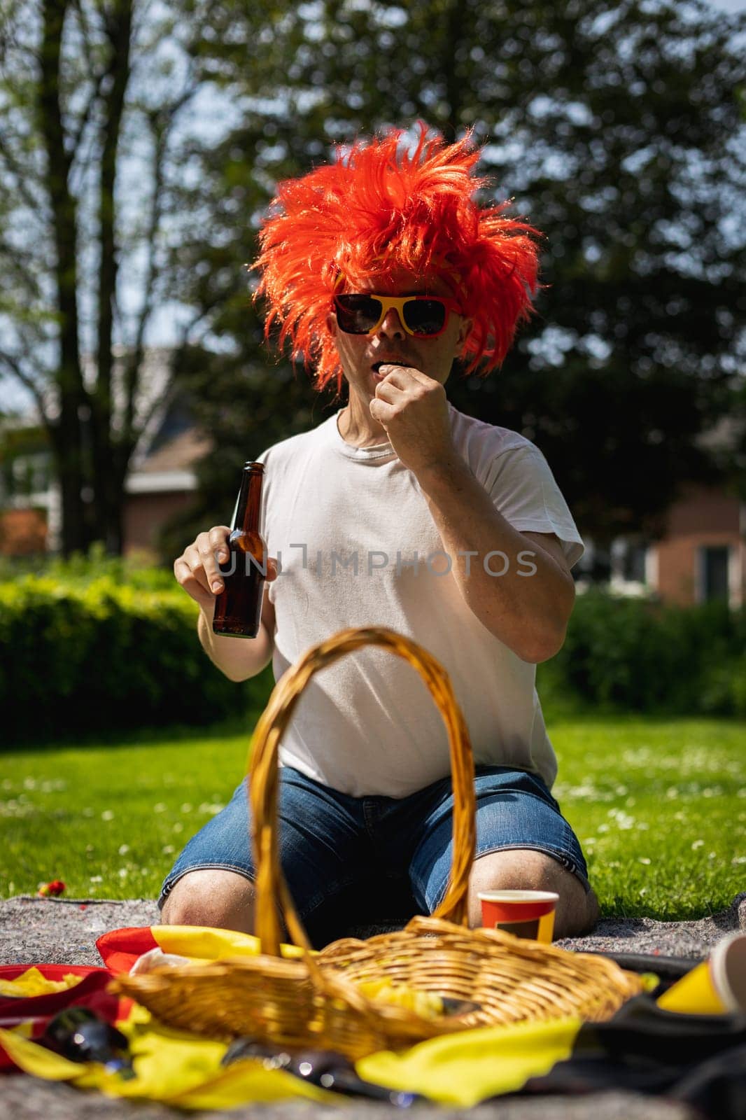 Portrait of a handsome young caucasian man in sunglasses, red wig drinking beer and eating chips celebrating belgium day at a picnic in a city park on a sunny summer day, close up side view.
