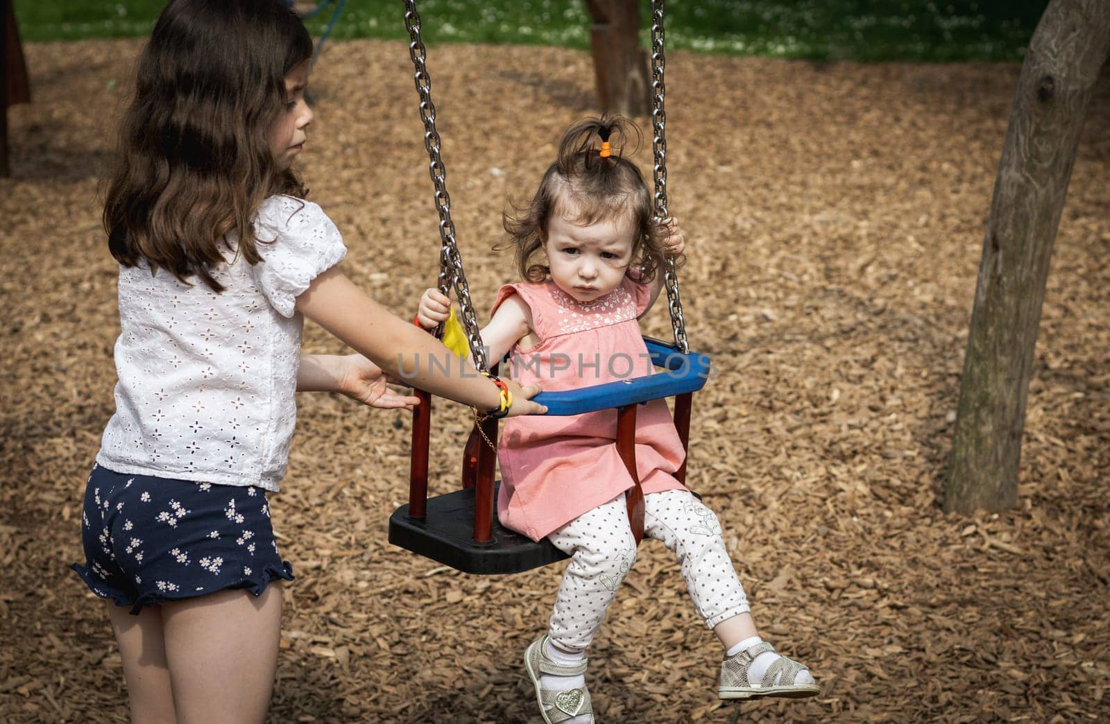 Portrait of a girl pushing her younger sister on a swing. by Nataliya