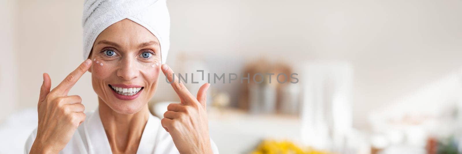 Mature beauty concept. Charming middle aged woman applying face cream under her eyes and smiling at camera. Lady taking care of her skin, pampering herself, panorama with copy space