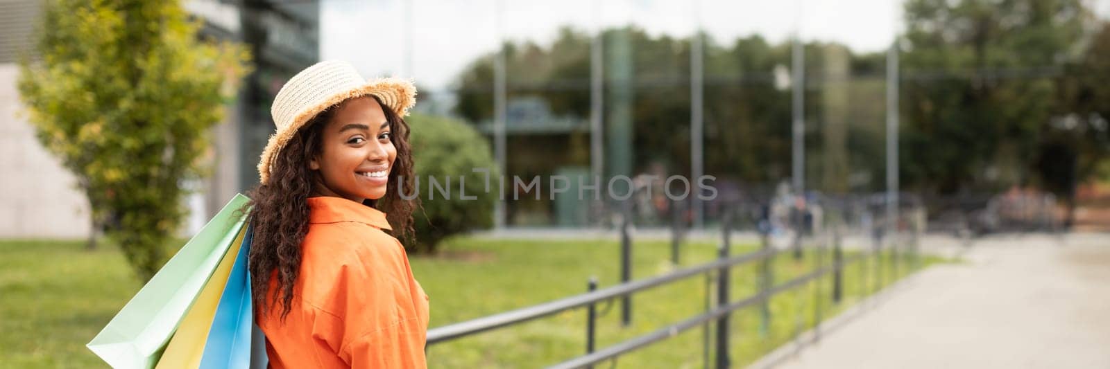 Cheerful millennial african american woman in casual and hat with many bags with purchases, looking at camera in city street. Urban walk, black friday sale, lifestyle and discount, copy space