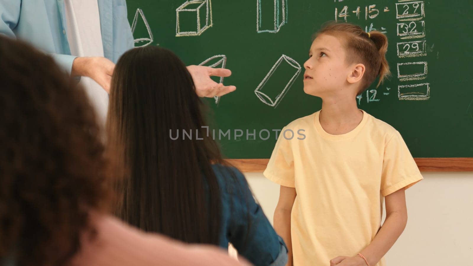 Professional teacher talking and explain idea to asian child during class. Caucasian student listening instructor while standing in front of classroom with blackboard math lesson written. Pedagogy.