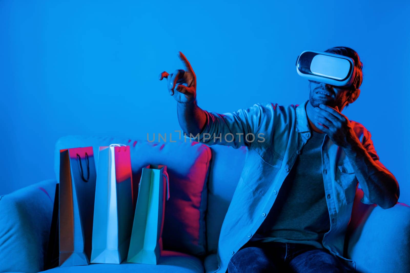 Caucasian man with vr glass posing pointing finger while sitting at sofa with shopping bags. Person selecting and doing shopping online by using VR technology and virtual goggle at home. Deviation.