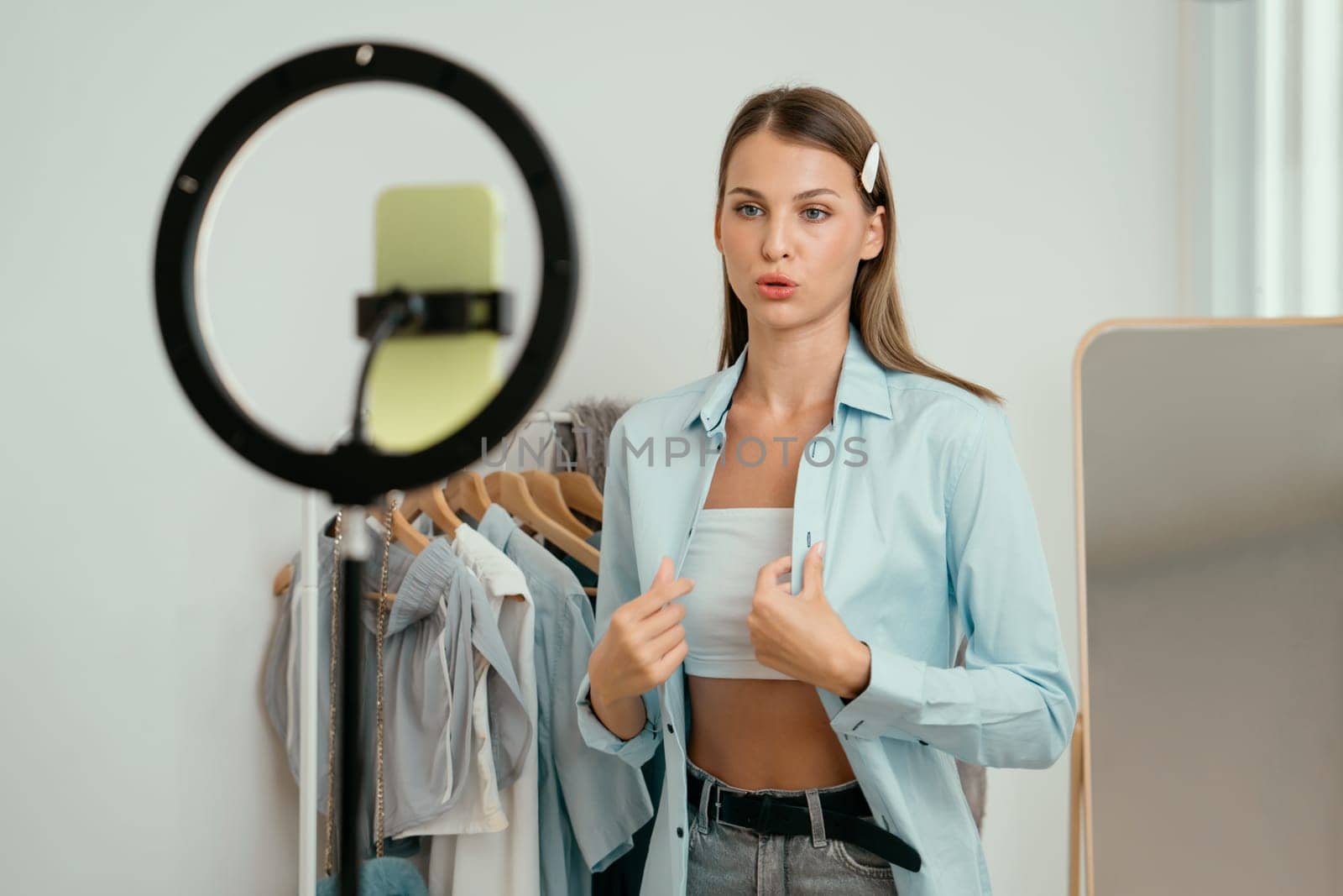 Young woman making fashion video content for social media. Blogger smiles to camera using selfie stick and light ring while making persuasive online clothing sell to audience or followers. Unveiling