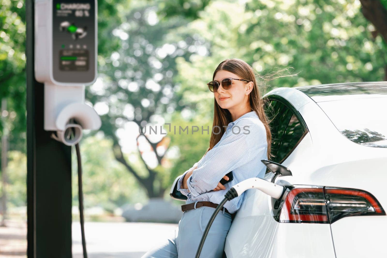 Young woman recharging battery for electric car during road trip travel EV car in natural forest or national park. Eco friendly travel during vacation and holiday. Exalt