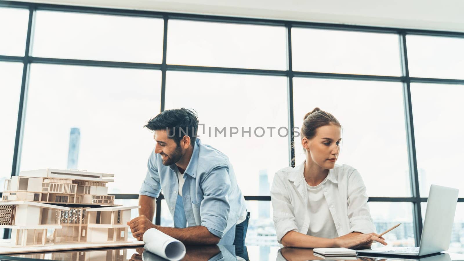 Young skilled project manager working by using laptop while caucasian engineer inspect house mode, using project plan at modern office or architect studio. Civil engineering, creative design. Tracery.