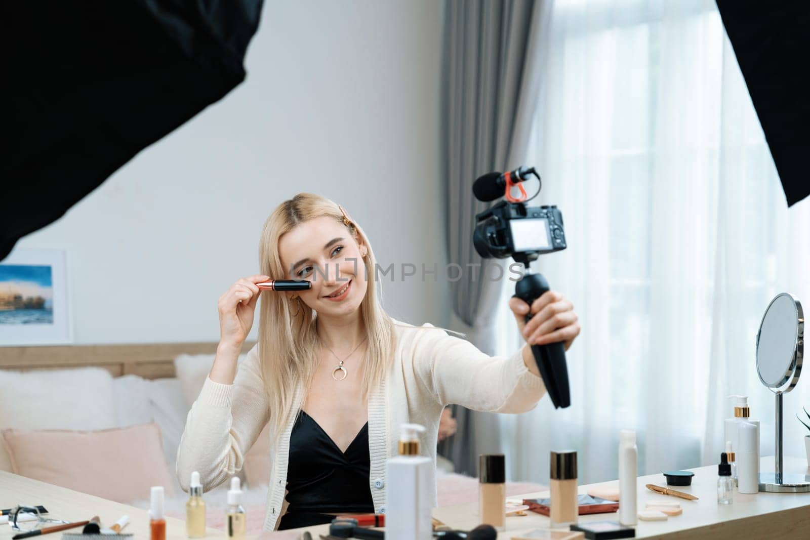 Young woman making beauty and cosmetic tutorial video content for social media. Beauty blogger smiles to camera while showing how to apply mascara to audience or followers. Blithe