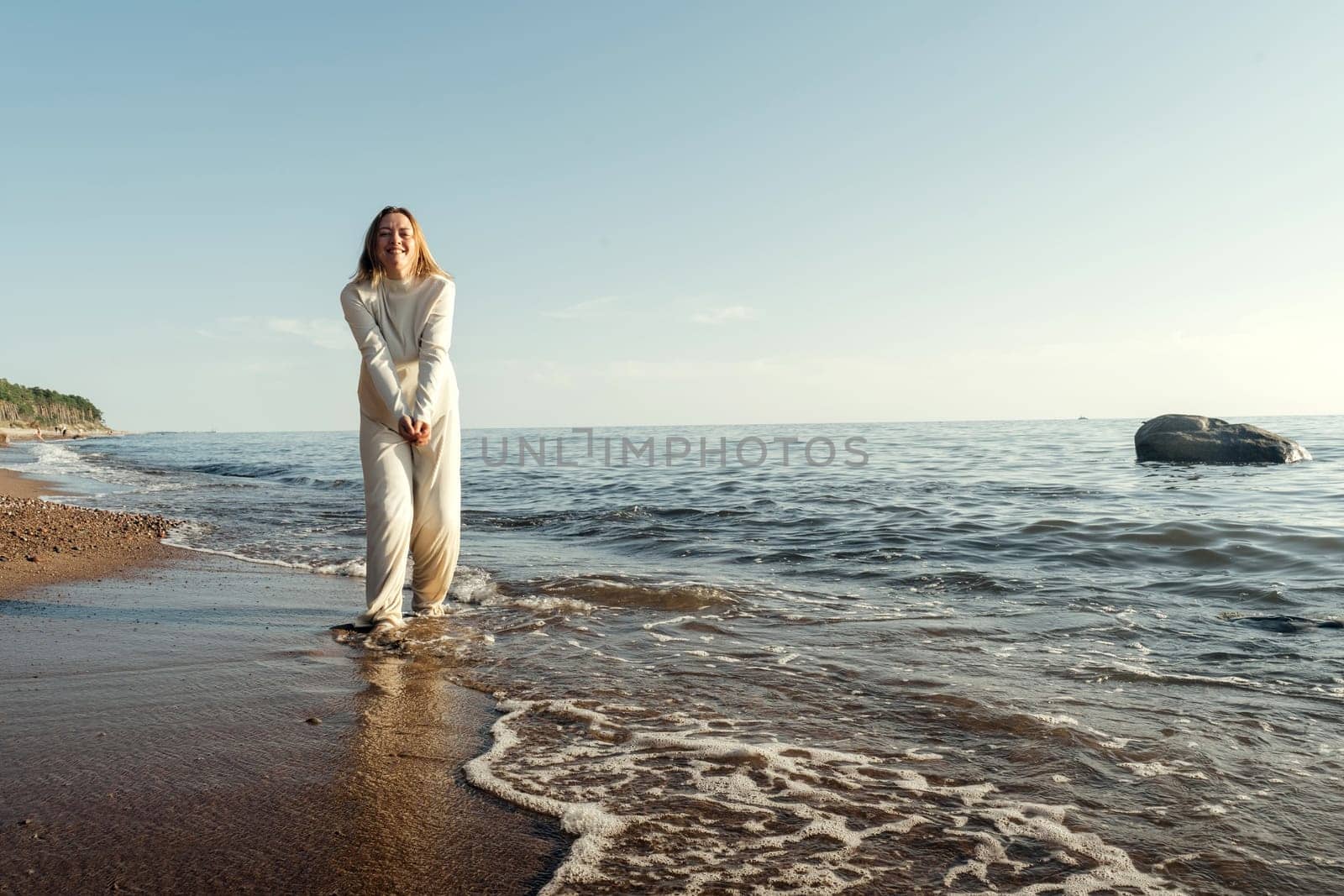 A woman is standing in the water at the beach, with waves gently lapping around her. She gazes out towards the horizon under the clear blue sky.