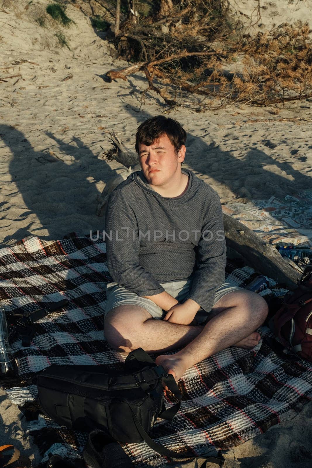 A man sits on a blanket on the sandy beach, gazing out at the ocean. The sun shines brightly overhead, casting a warm glow on the scene.